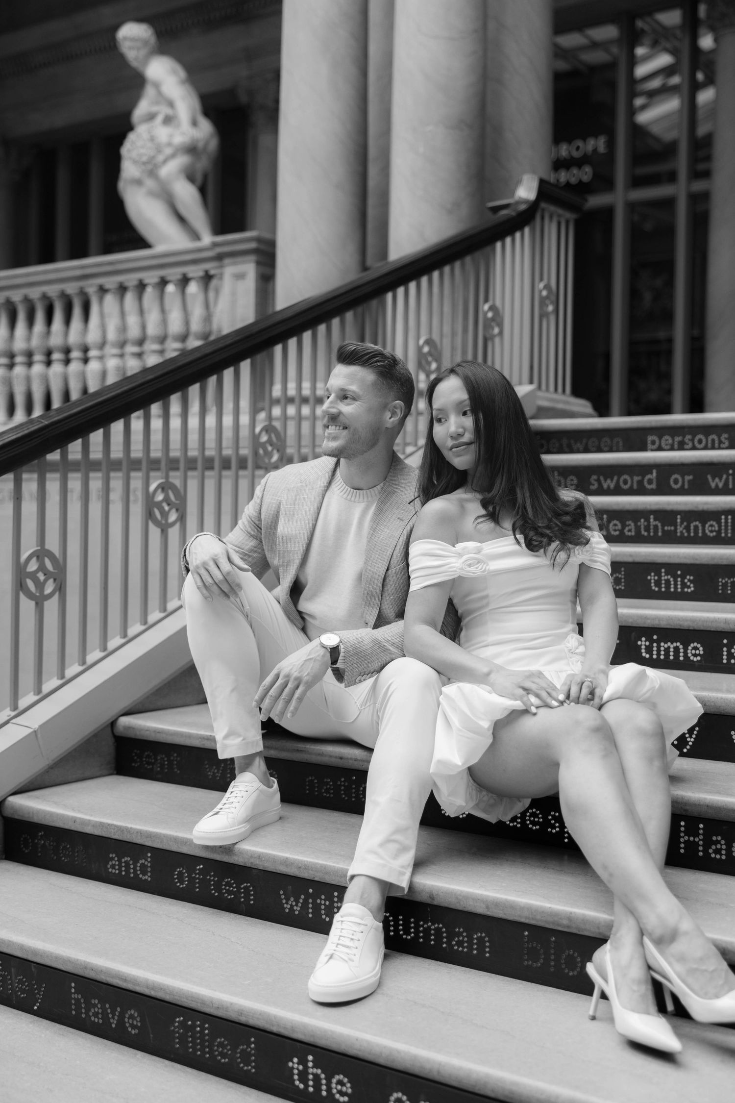 Black and white photo of a couple sitting on decorative steps outside a classical building with a statue.