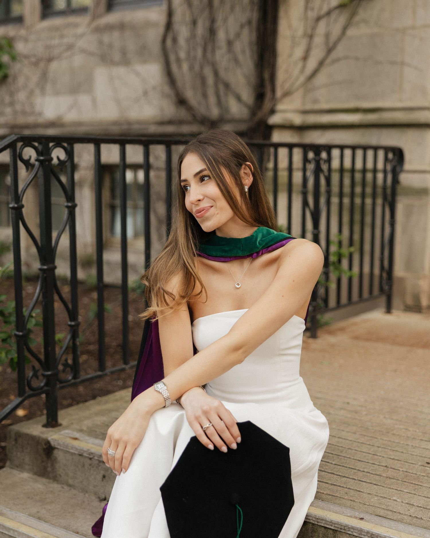Young woman in white dress with green and purple scarf sits on steps near iron fence outdoors.