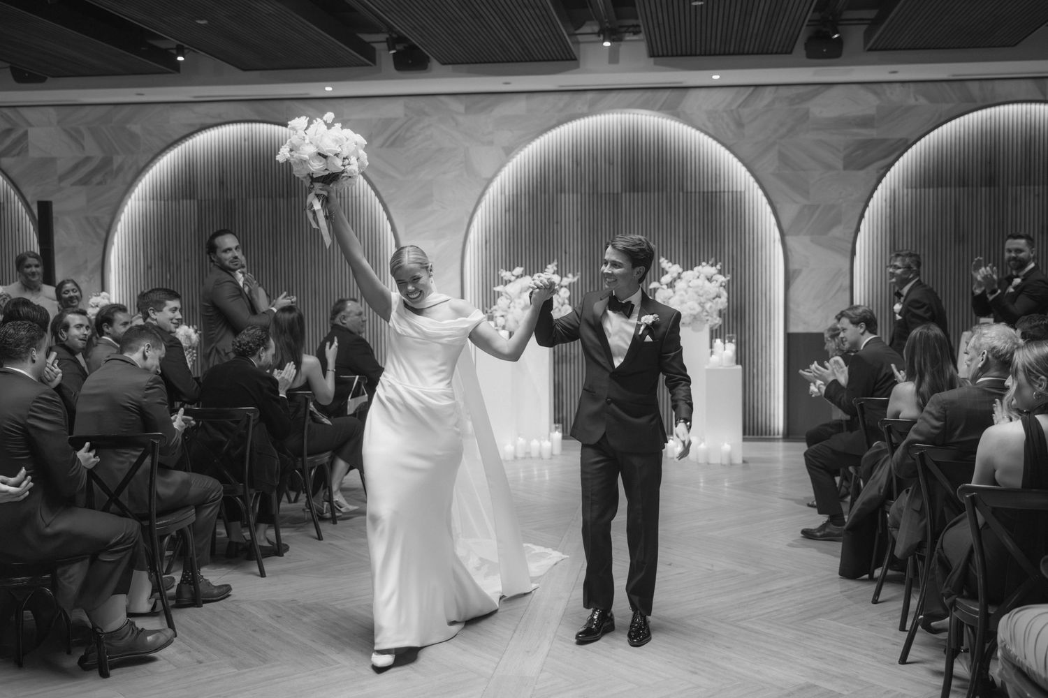 Bride and groom triumphantly raise bouquets while exiting wedding ceremony in black and white.