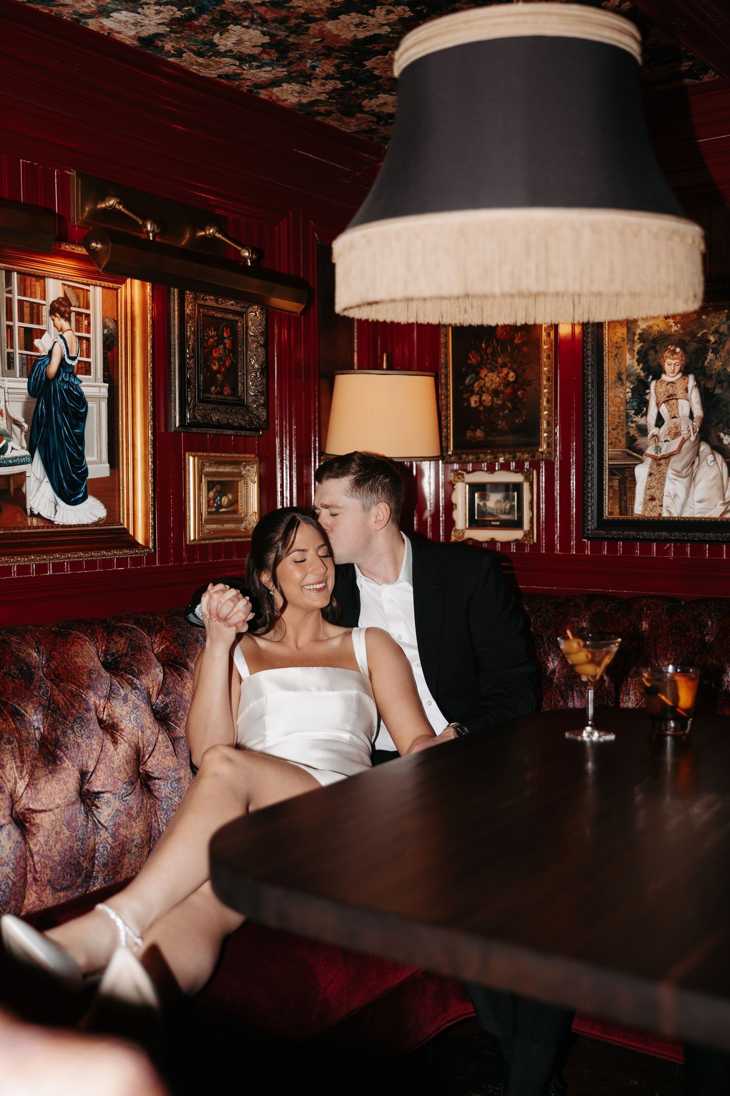 A romantic scene unfolds on a tufted red leather banquette beneath vintage artwork and a large fringed lamp.