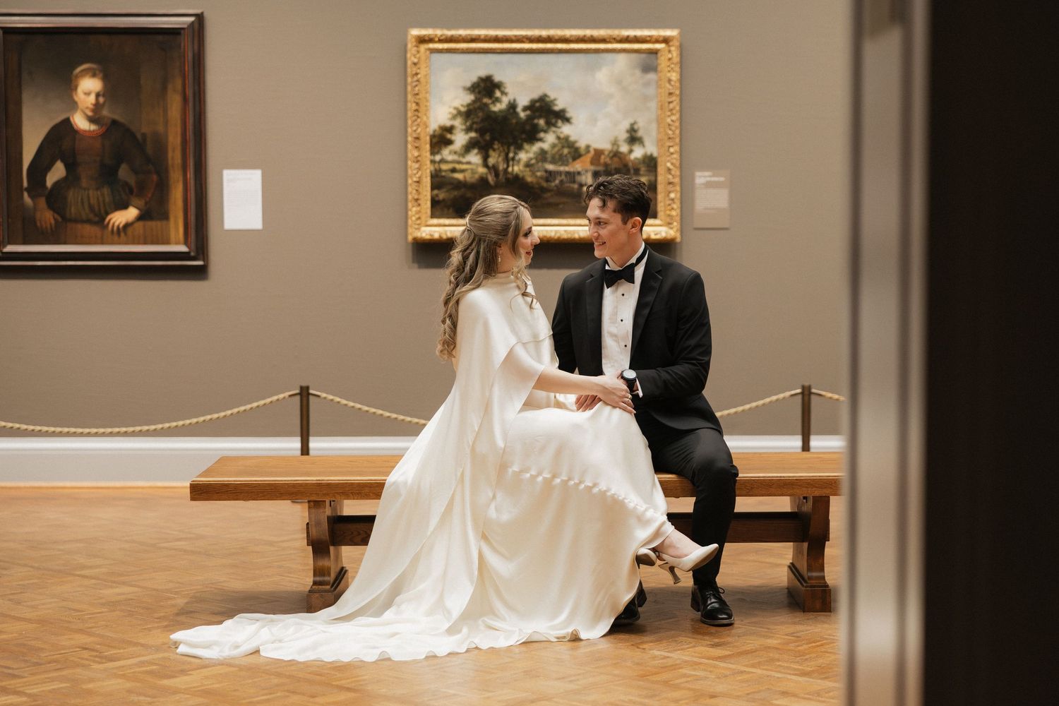 Bride and groom sitting on museum bench in wedding attire, surrounded by classic paintings in an art gallery.