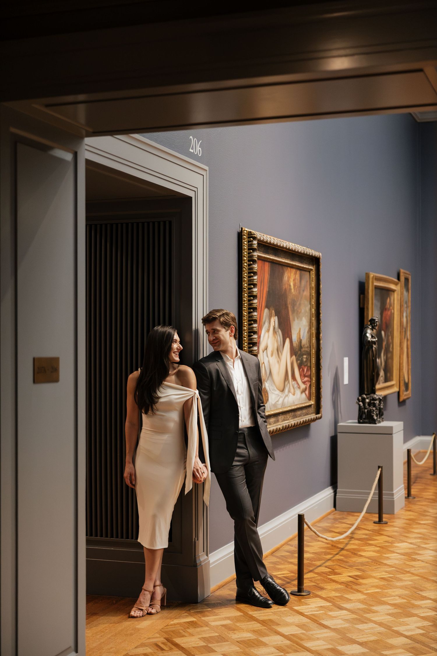 Elegant couple in evening attire smiling in a museum gallery with framed oil paintings, velvet ropes and herringbone wood floors.