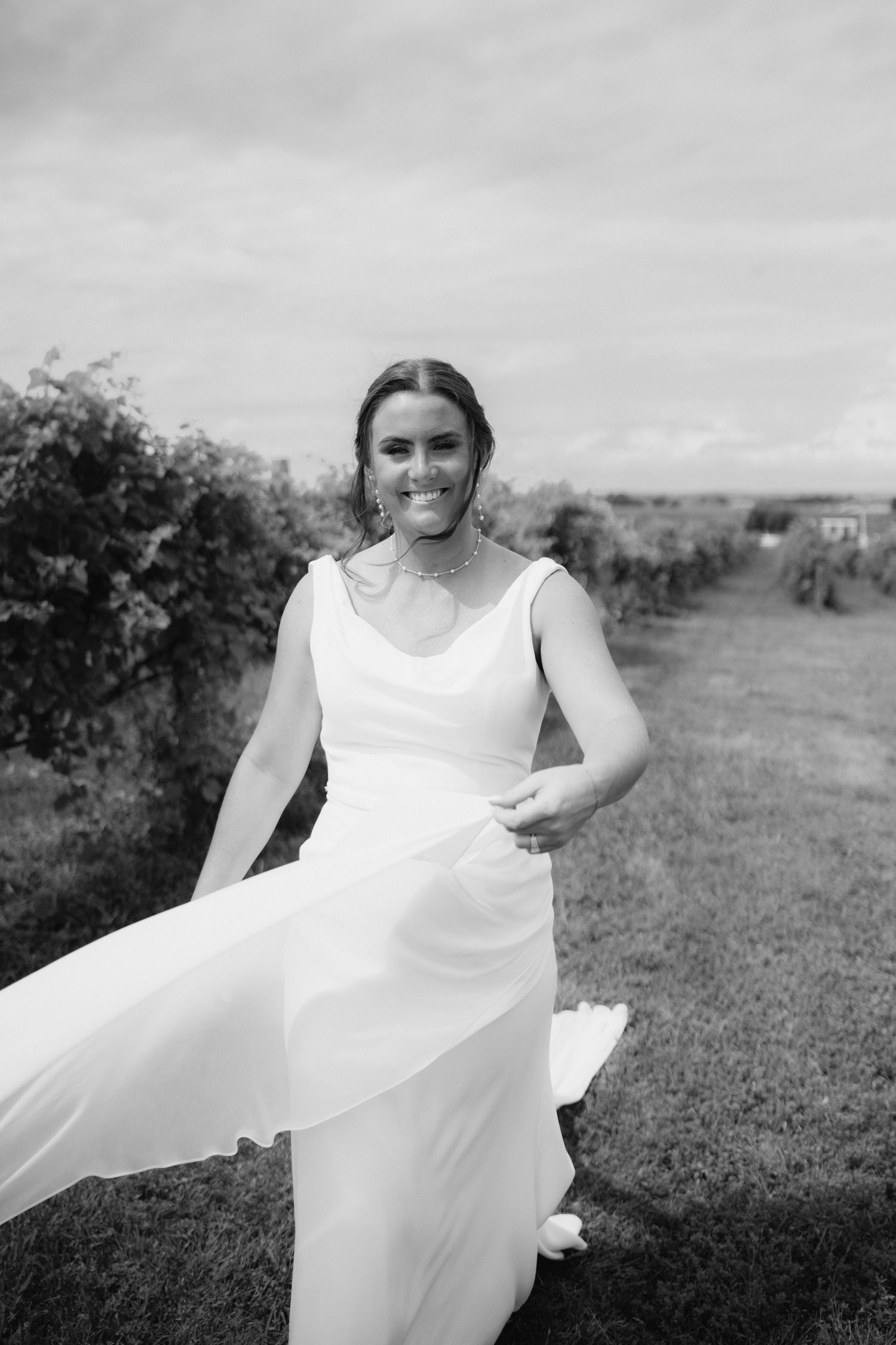 A bride in a flowing white dress twirls in a vineyard on an overcast day.