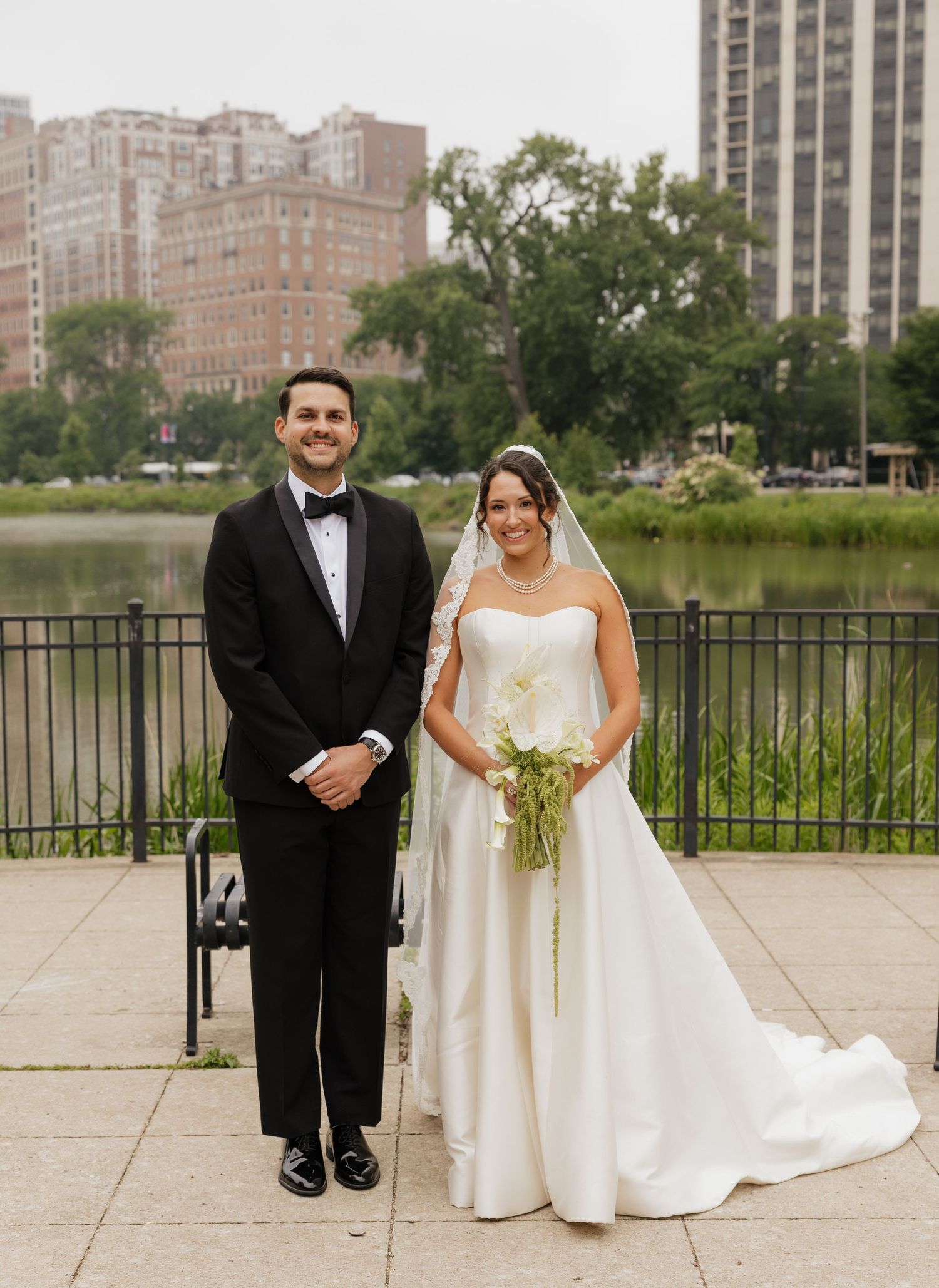 Bride and groom posing by a lakeside park with city buildings in the background on their wedding day.