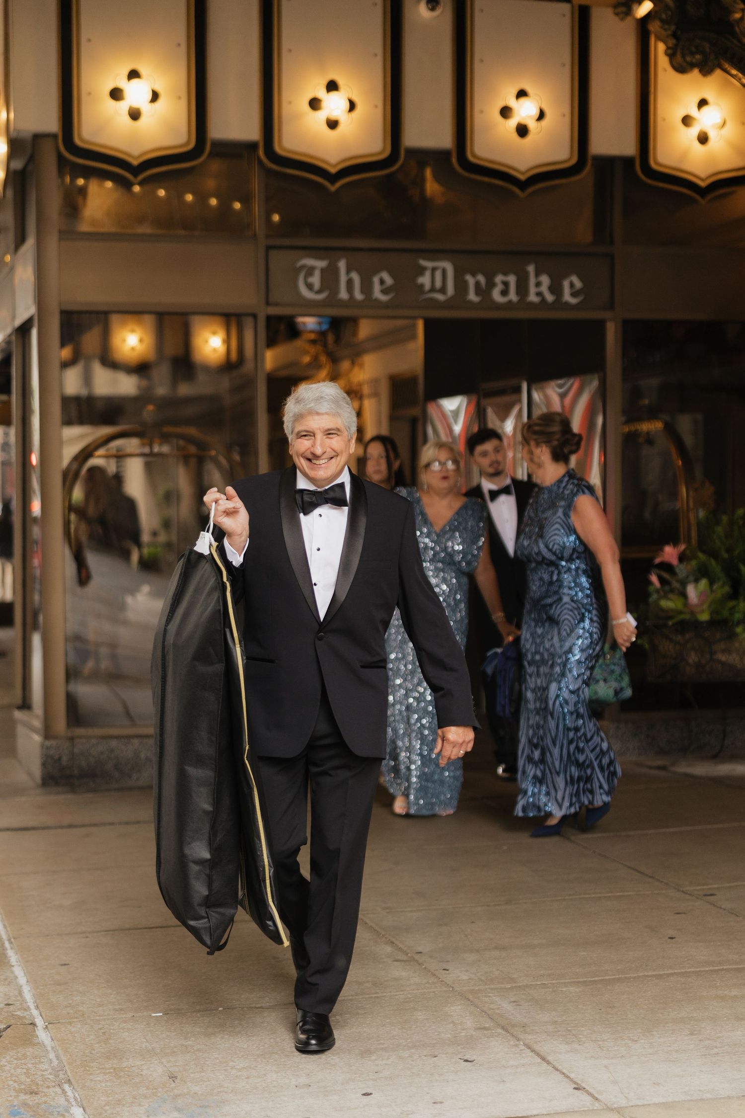 Elegant man in tuxedo exits The Drake Hotel carrying a garment bag, with formally dressed guests following behind.