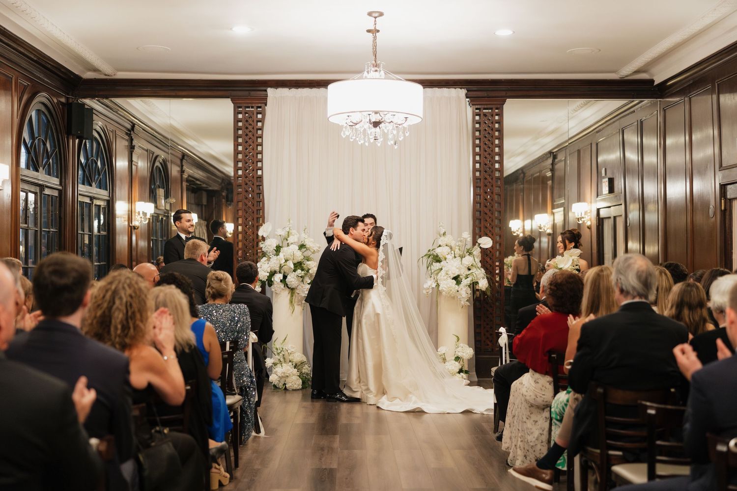 Bride and groom share first kiss at elegant indoor wedding ceremony with white floral arch and chandelier.