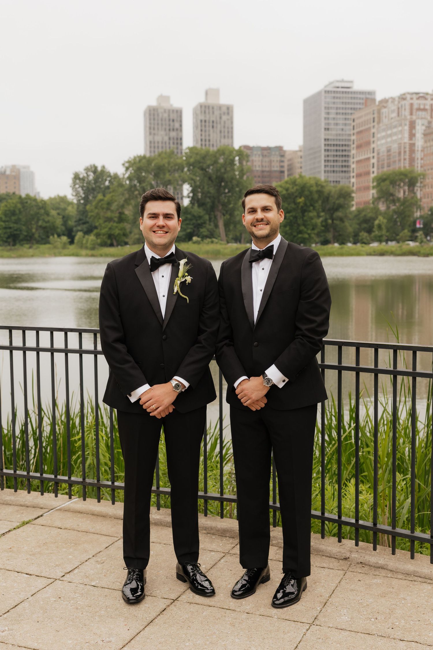 Two grooms in black tuxedos with bow ties pose by a lakeside railing with a city skyline backdrop.