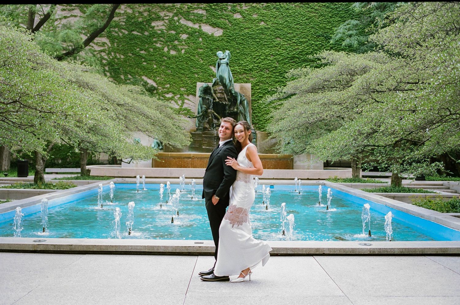Bride and groom posing by a fountain with ivy-covered walls and bronze sculpture at an elegant outdoor venue.
