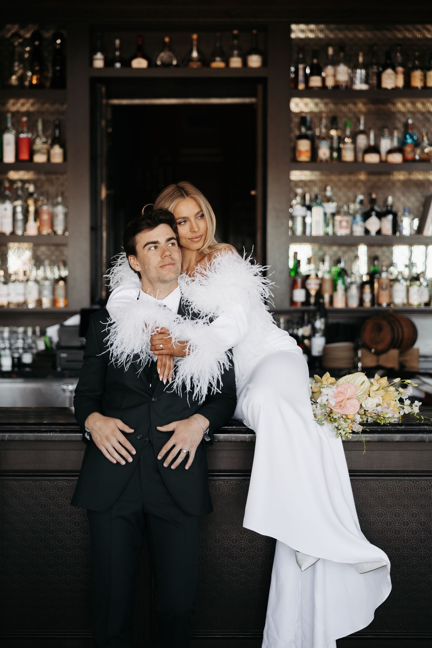 Stylish couple poses playfully at a modern bar counter with bottles and glassware in the background.