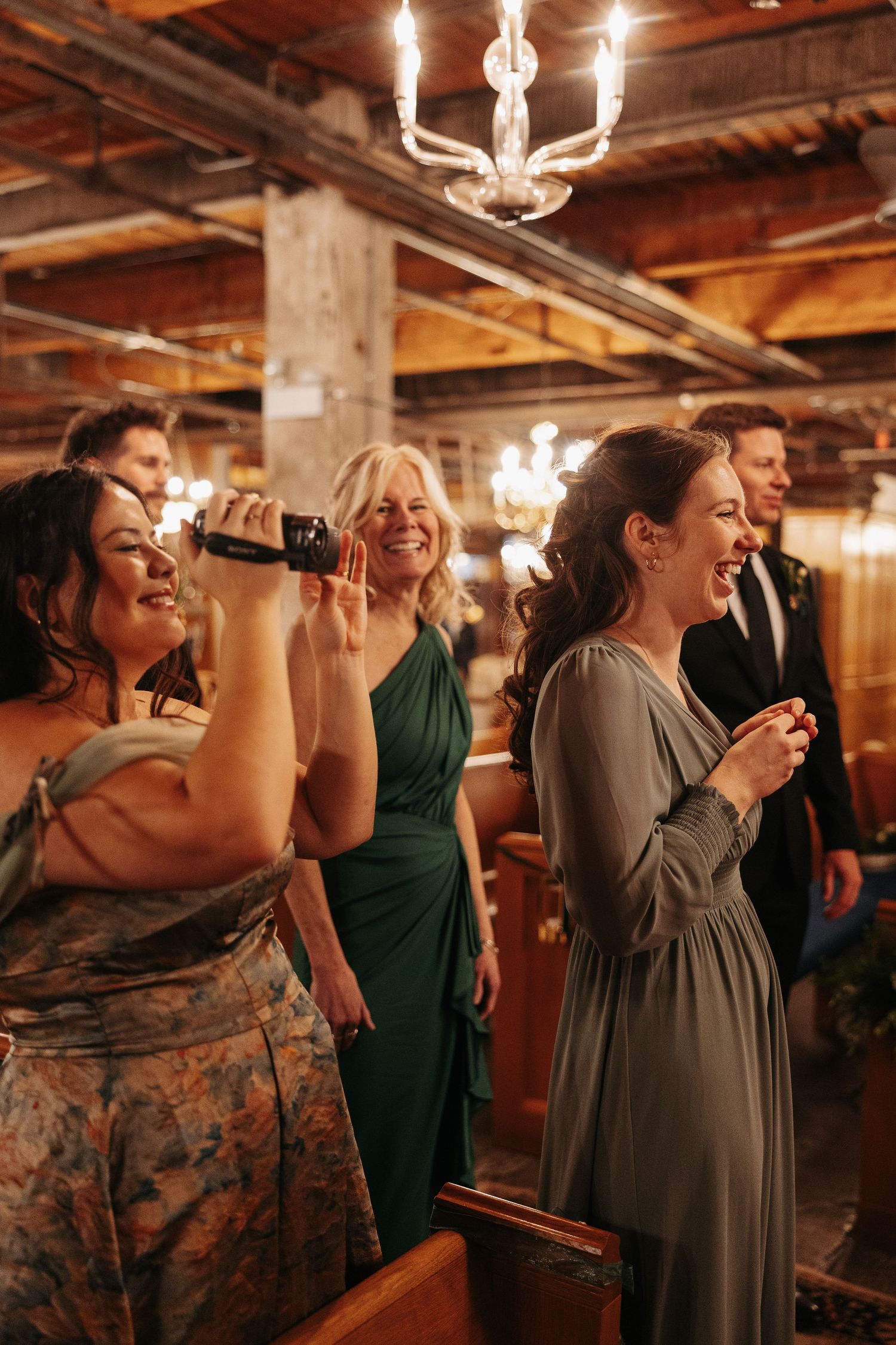 Wedding party guests enjoy drinks and dancing in a rustic venue with wooden beams and chandeliers.
