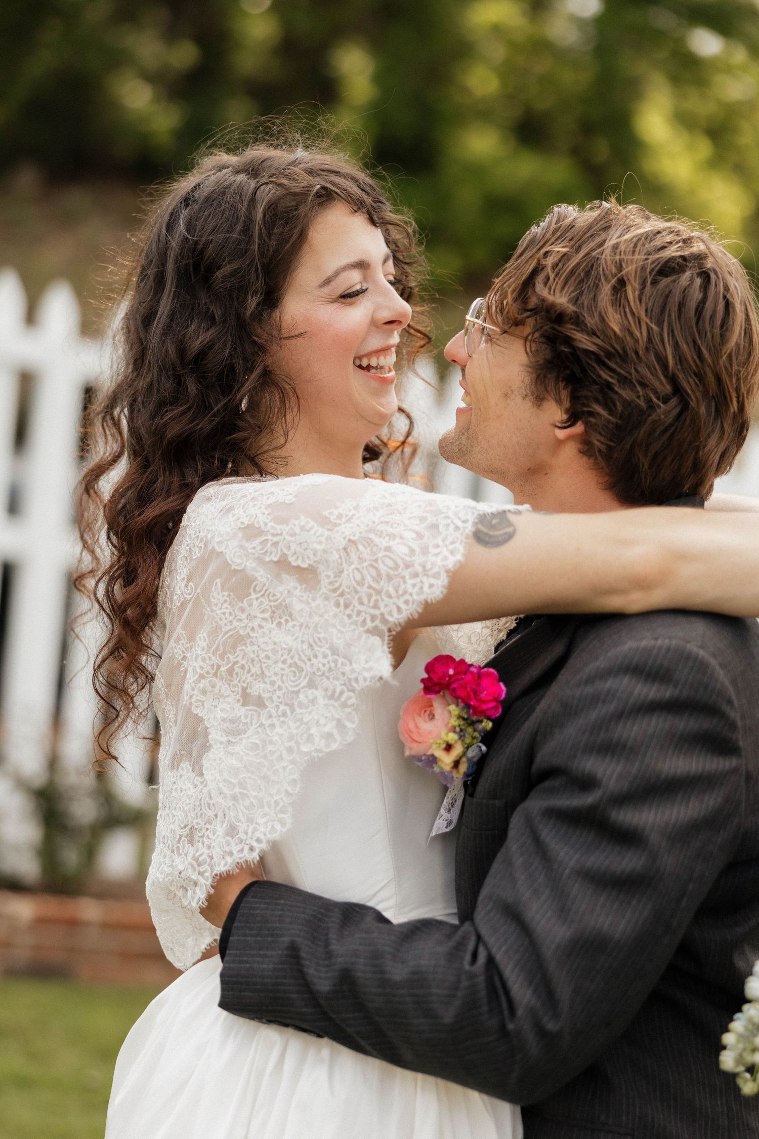 Joyful bride in lace gown embracing groom in gray suit outdoors at a wedding ceremony.