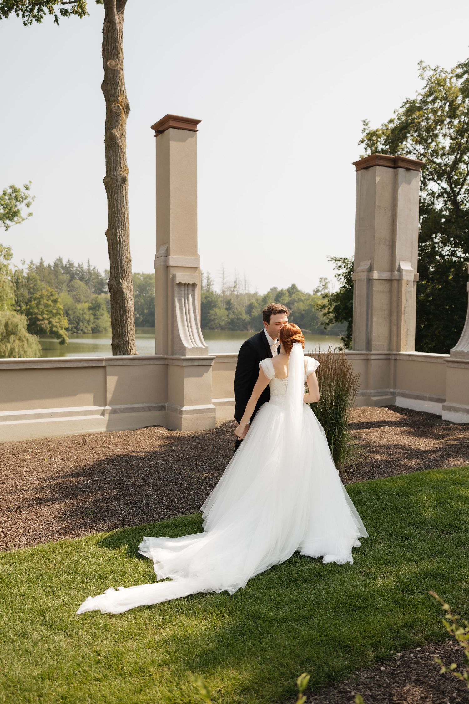 Bride and groom share a romantic kiss outdoors near elegant stone columns with a serene lake view on their wedding day.