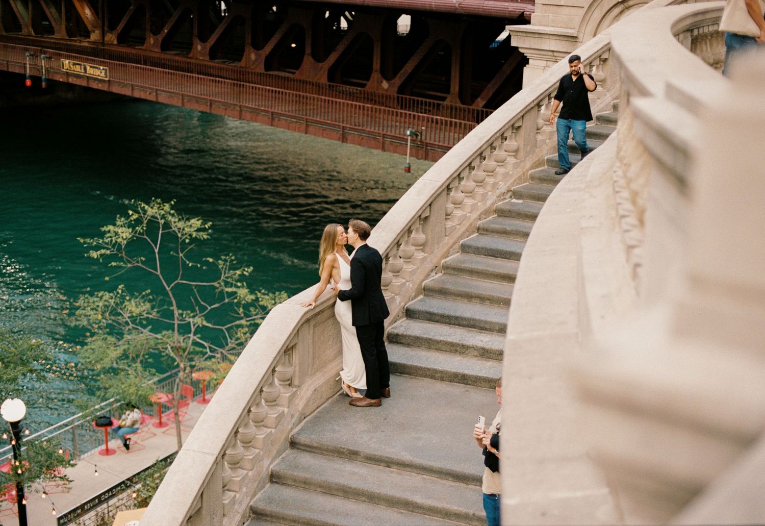 Couple sharing a romantic moment on ornate stone bridge stairs beside a turquoise river in an urban setting.