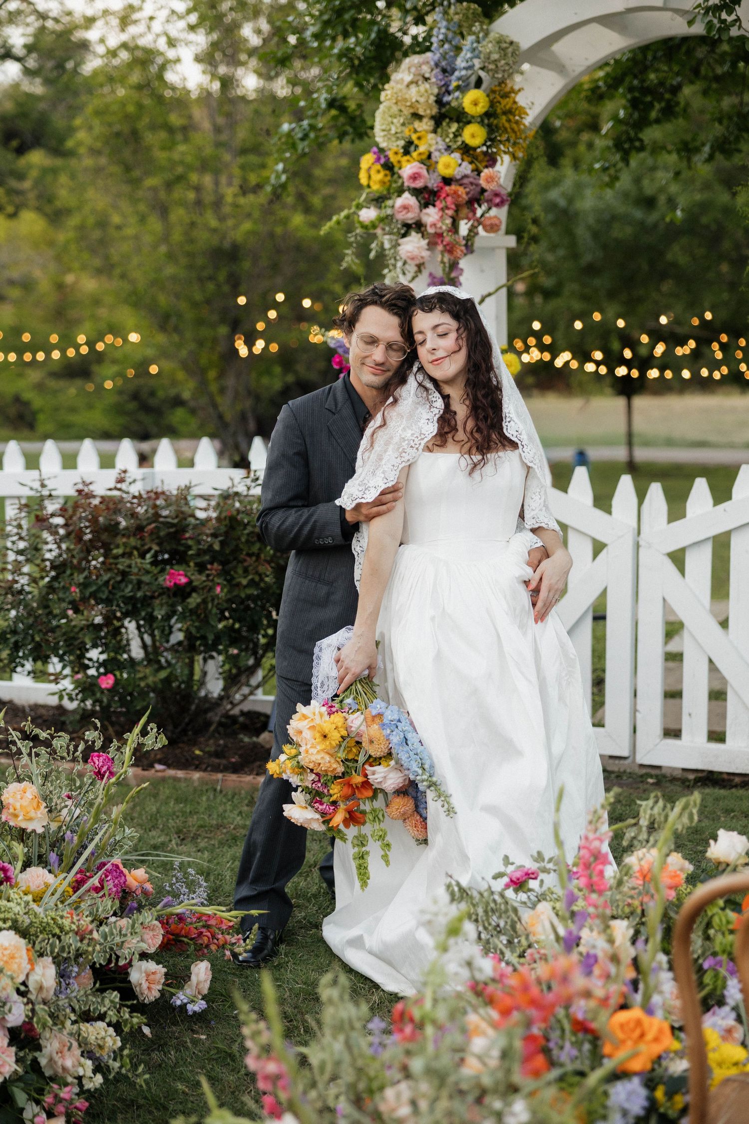 Bride and groom embrace under a floral arch with colorful blooms and string lights at an outdoor garden wedding.
