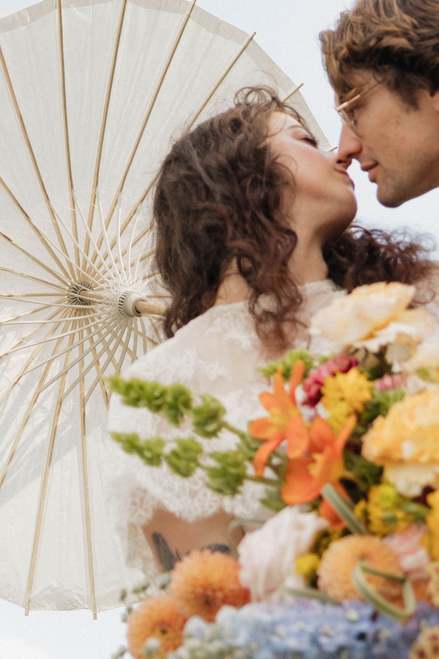 Romantic couple almost kissing, holding a white parasol and colorful floral bouquet at an outdoor wedding.