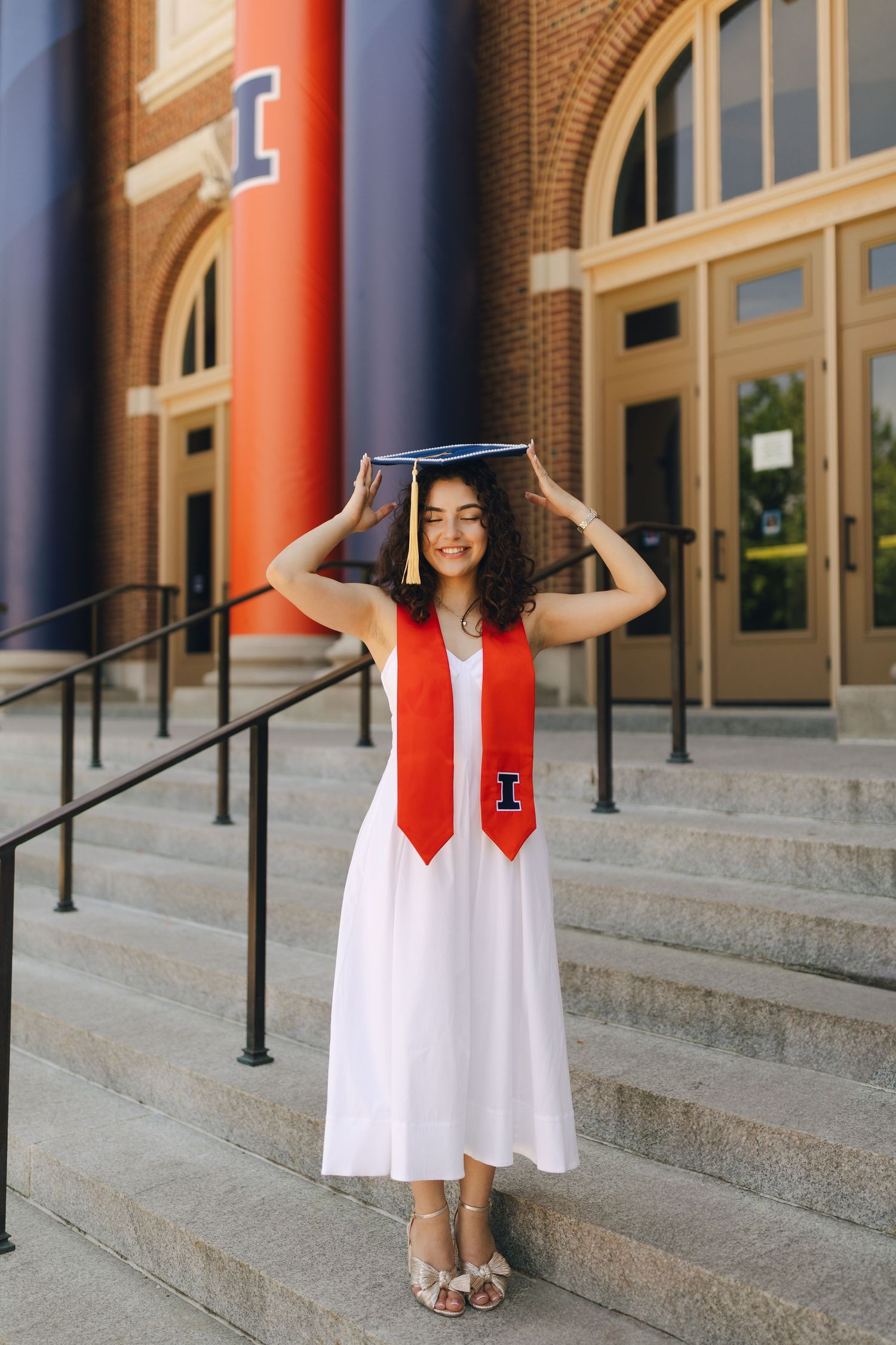 Graduate in white dress and red stole poses on campus steps with graduation cap and bright smile.