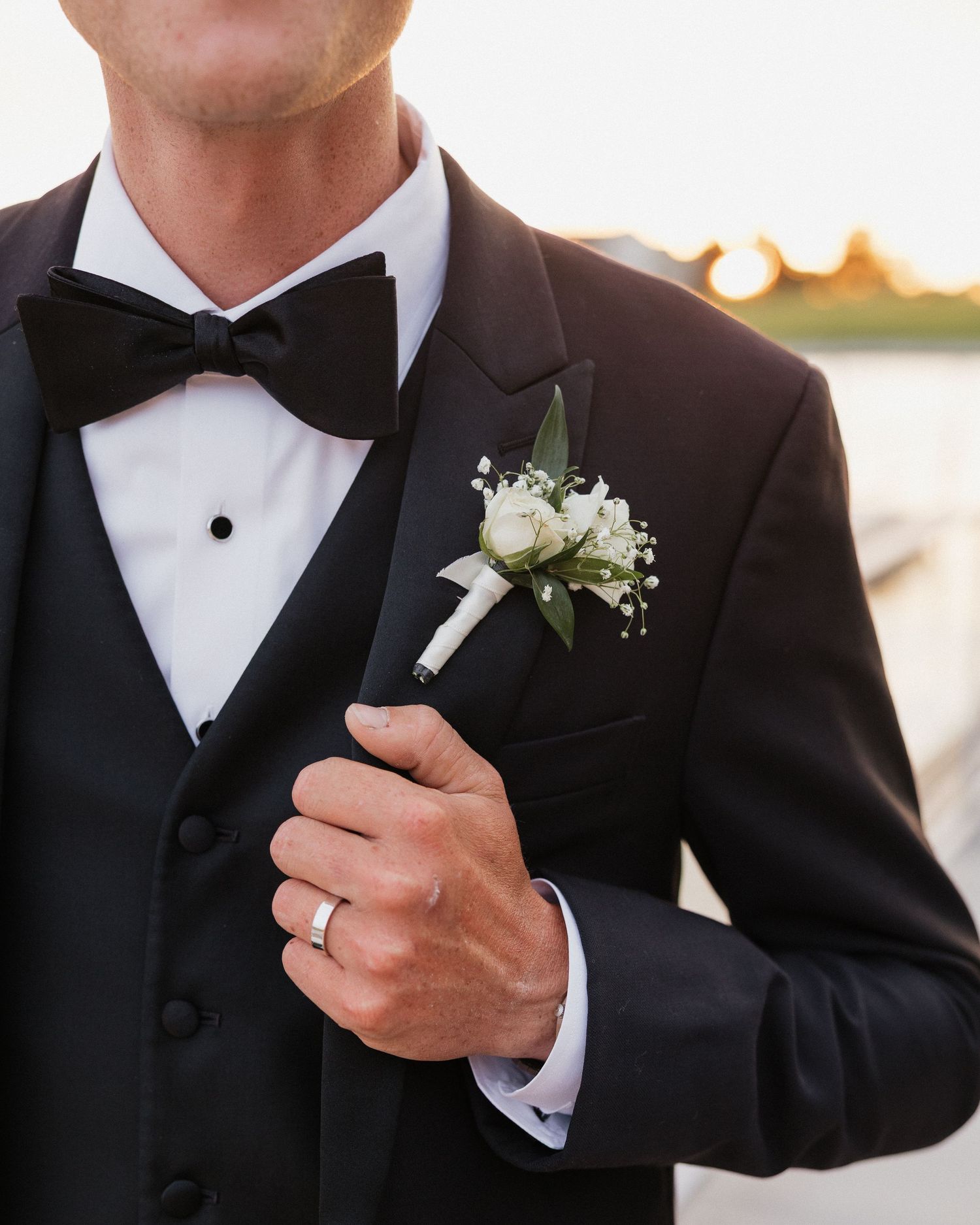 Groom in black tuxedo with bow tie and white rose boutonniere, wearing wedding ring, golden sunset backdrop.