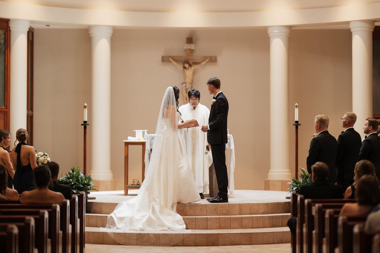 Bride and groom exchange vows at Catholic church altar with crucifix, guests seated in wooden pews during wedding ceremony.