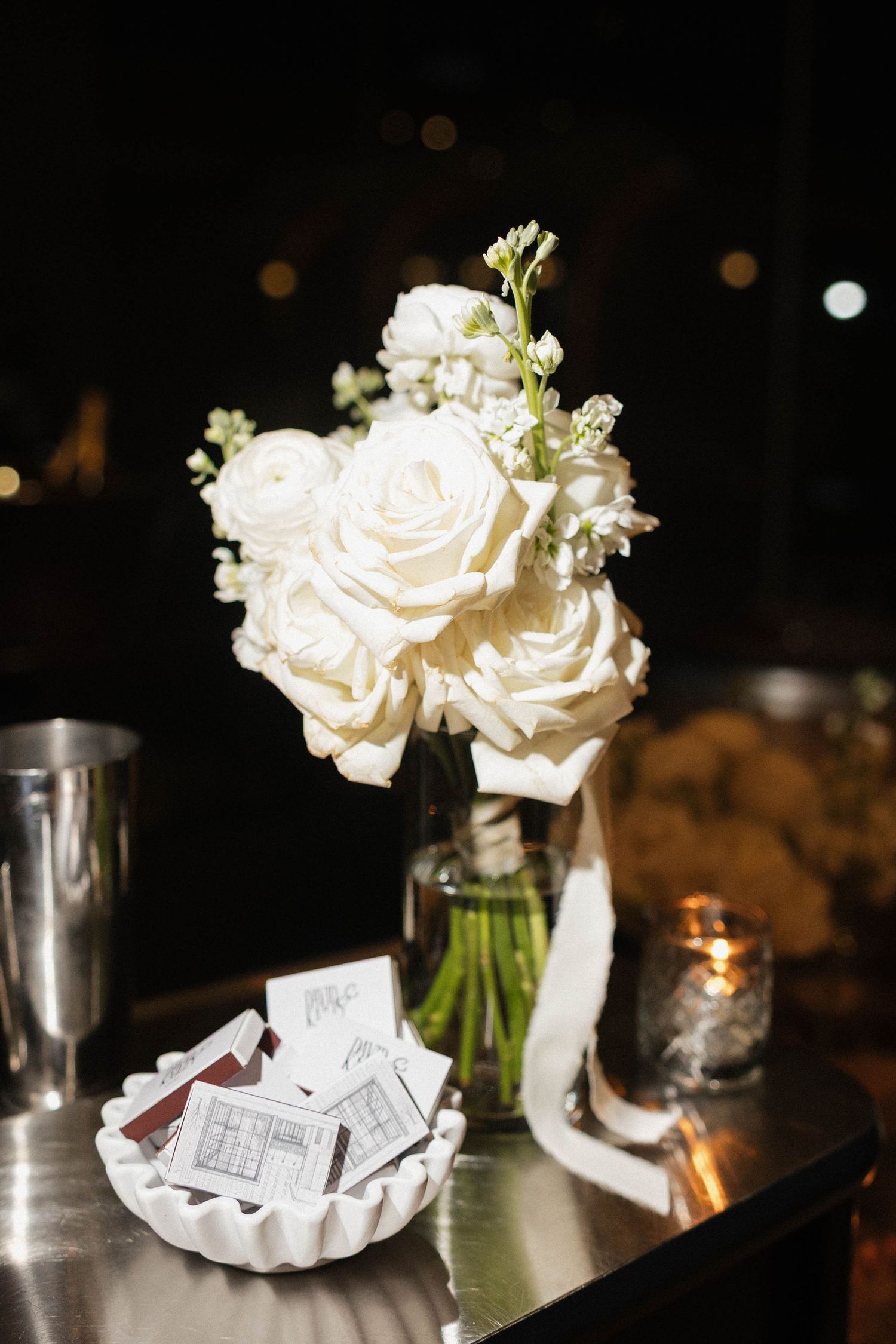 Elegant white rose bouquet in glass vase on bar table with matchbooks and candle at wedding reception.
