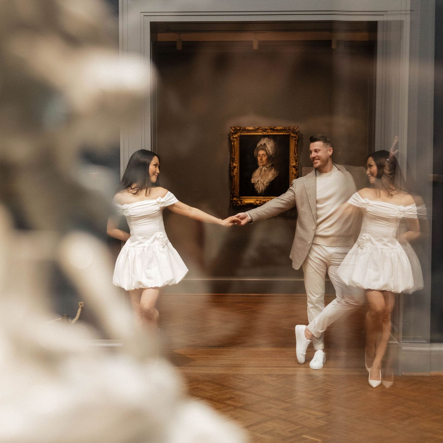 Couple in white outfits dancing joyfully in an elegant art museum gallery with a classic portrait painting backdrop.