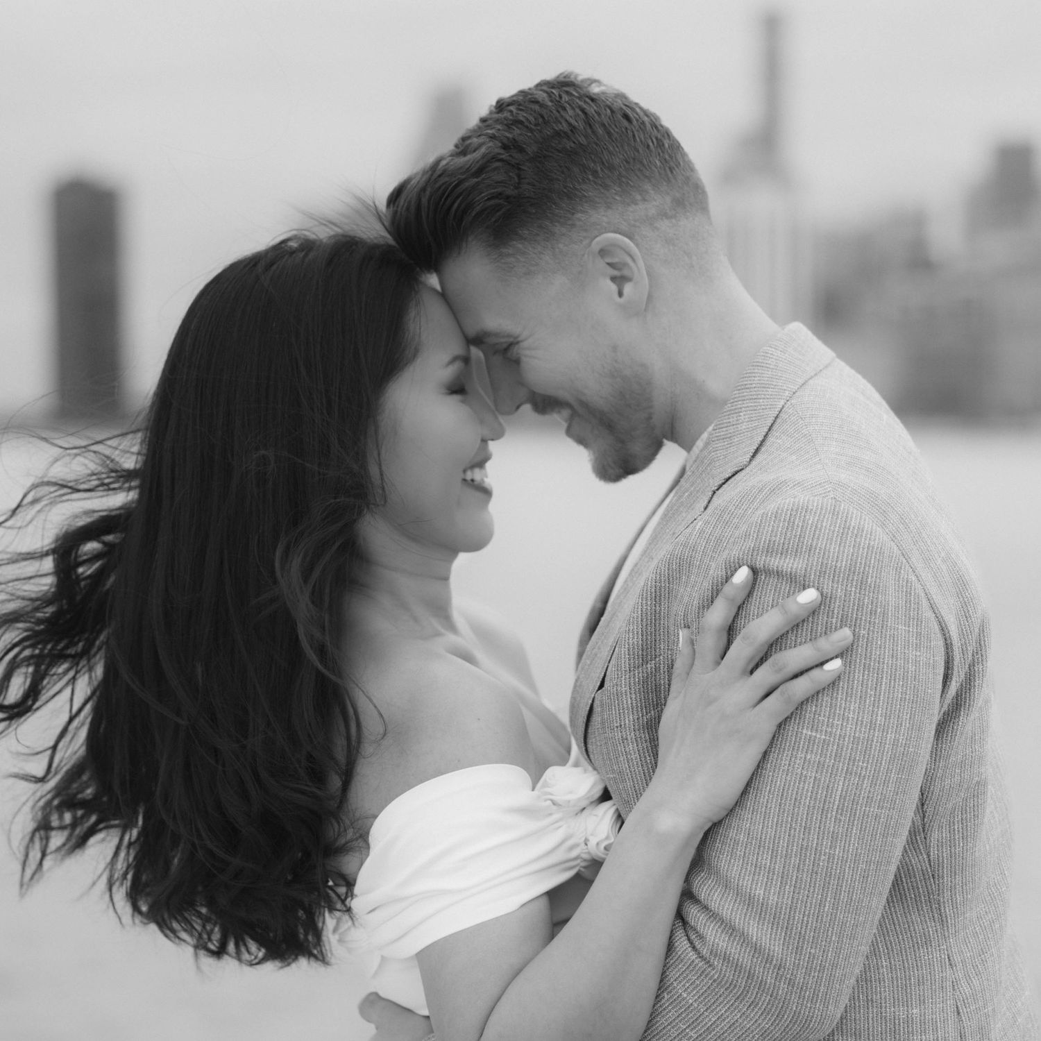 Black and white romantic photo of couple touching foreheads tenderly with city skyline softly blurred behind them.
