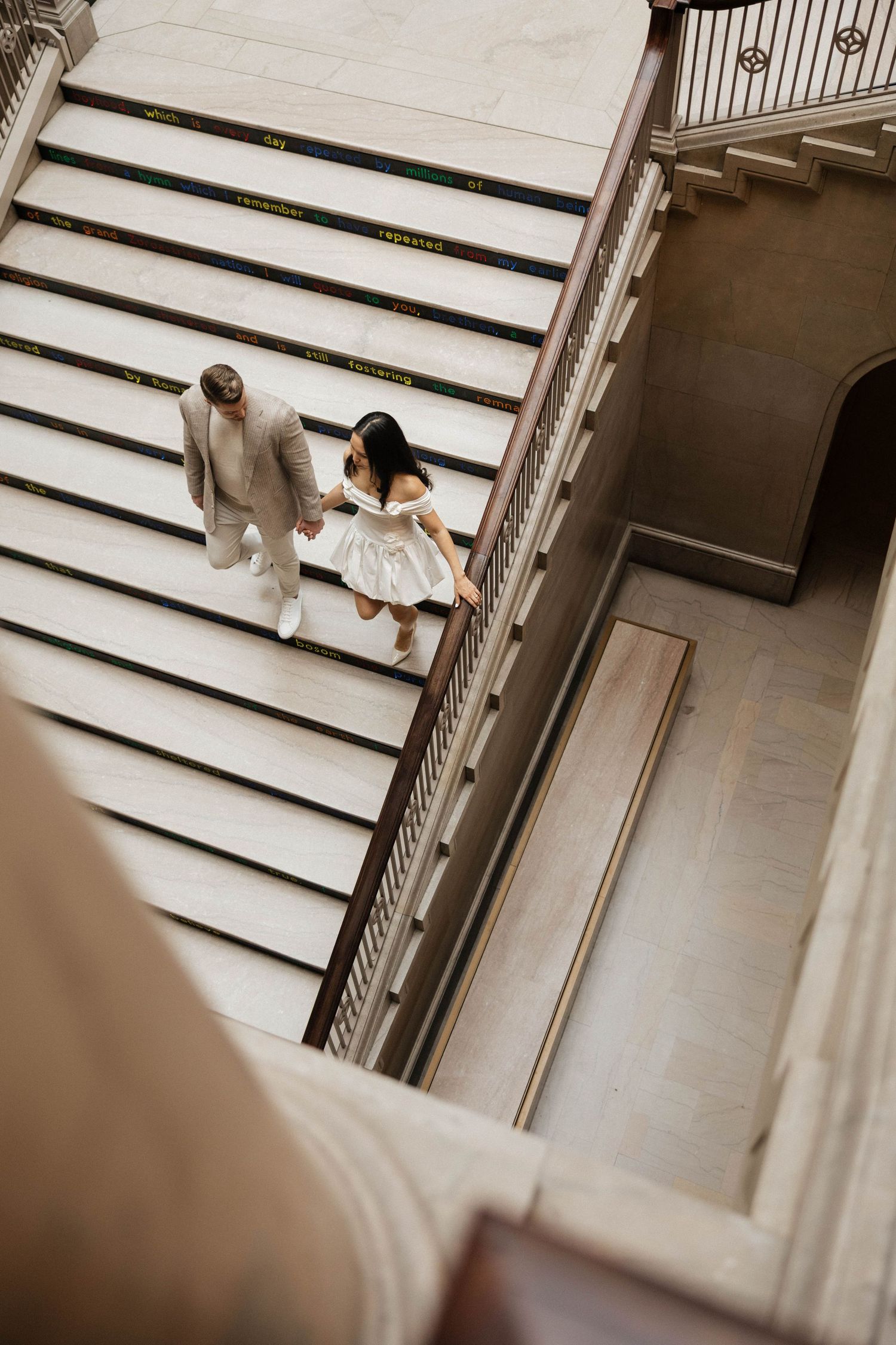 Aerial view of a couple holding hands walking up grand marble stairs, man in tan suit and woman in white dress.