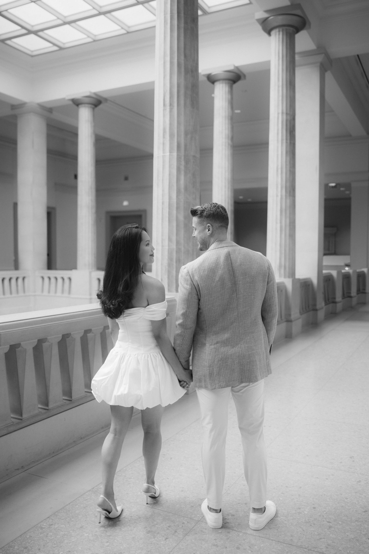 Black and white photo of a couple holding hands walking through an elegant columned hall.