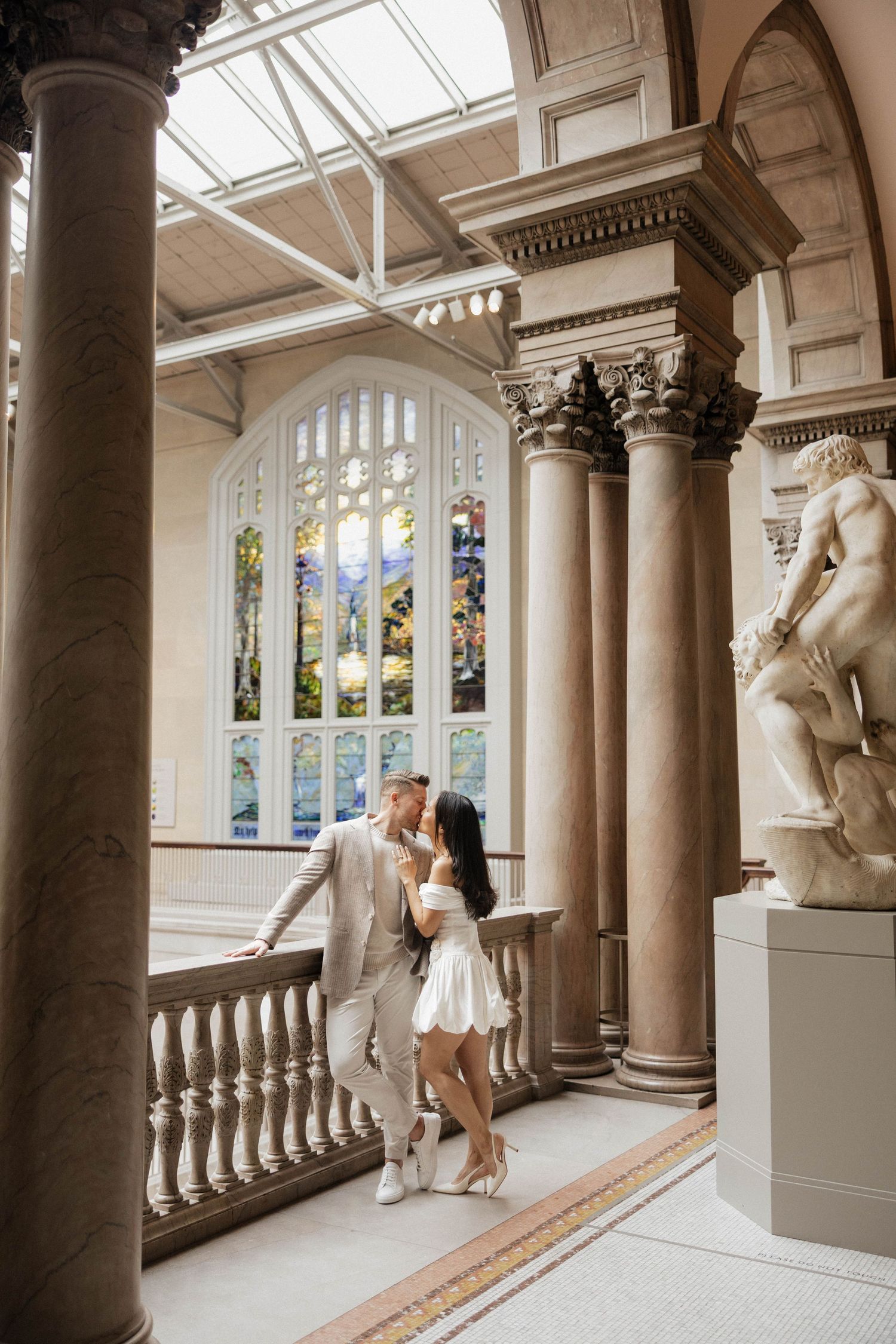 Couple sharing a romantic moment in an elegant museum hall with marble columns and stained glass windows.