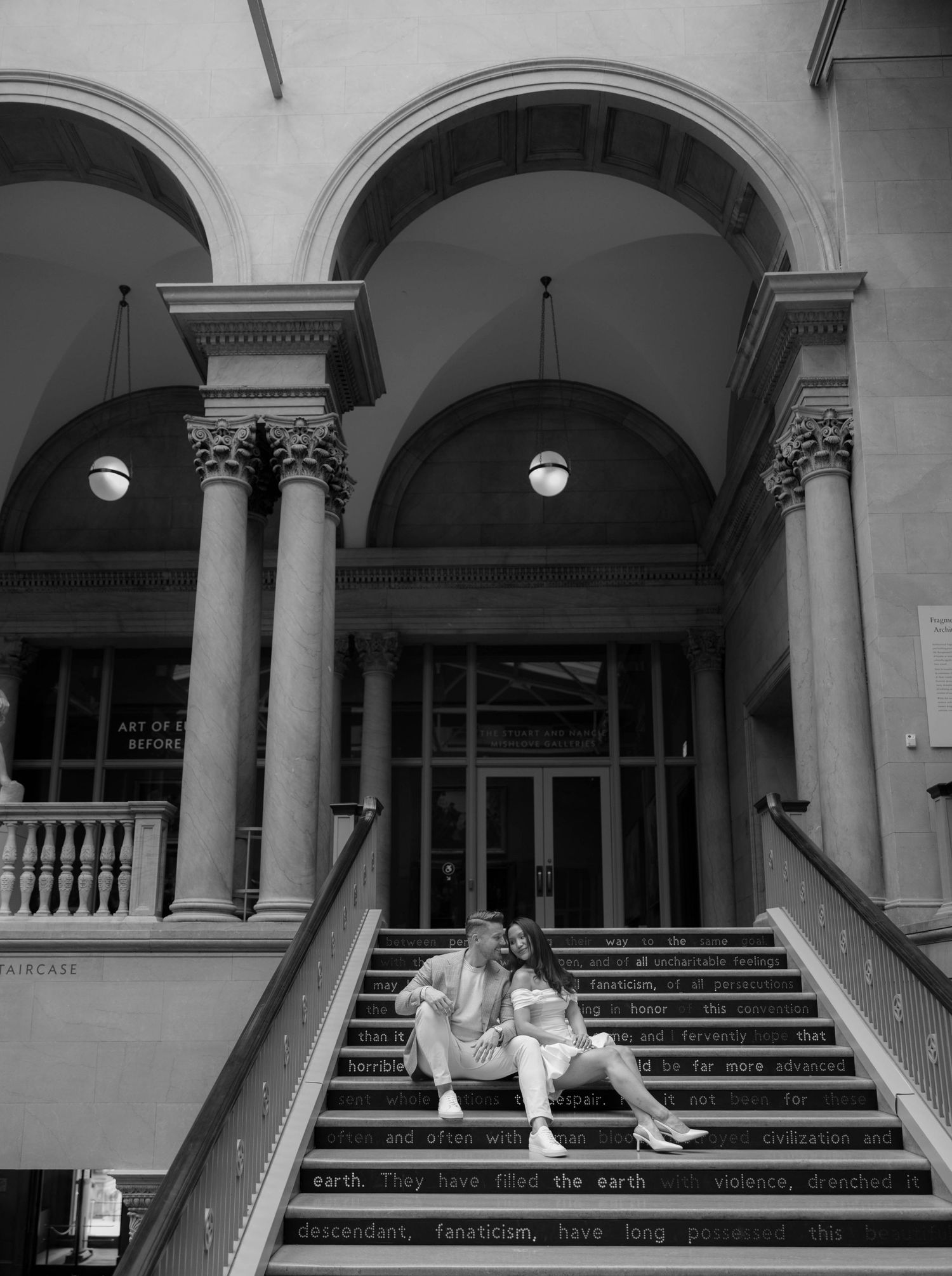 Couple sitting on text-inscribed steps of a grand neoclassical building with arched columns, black and white photo.