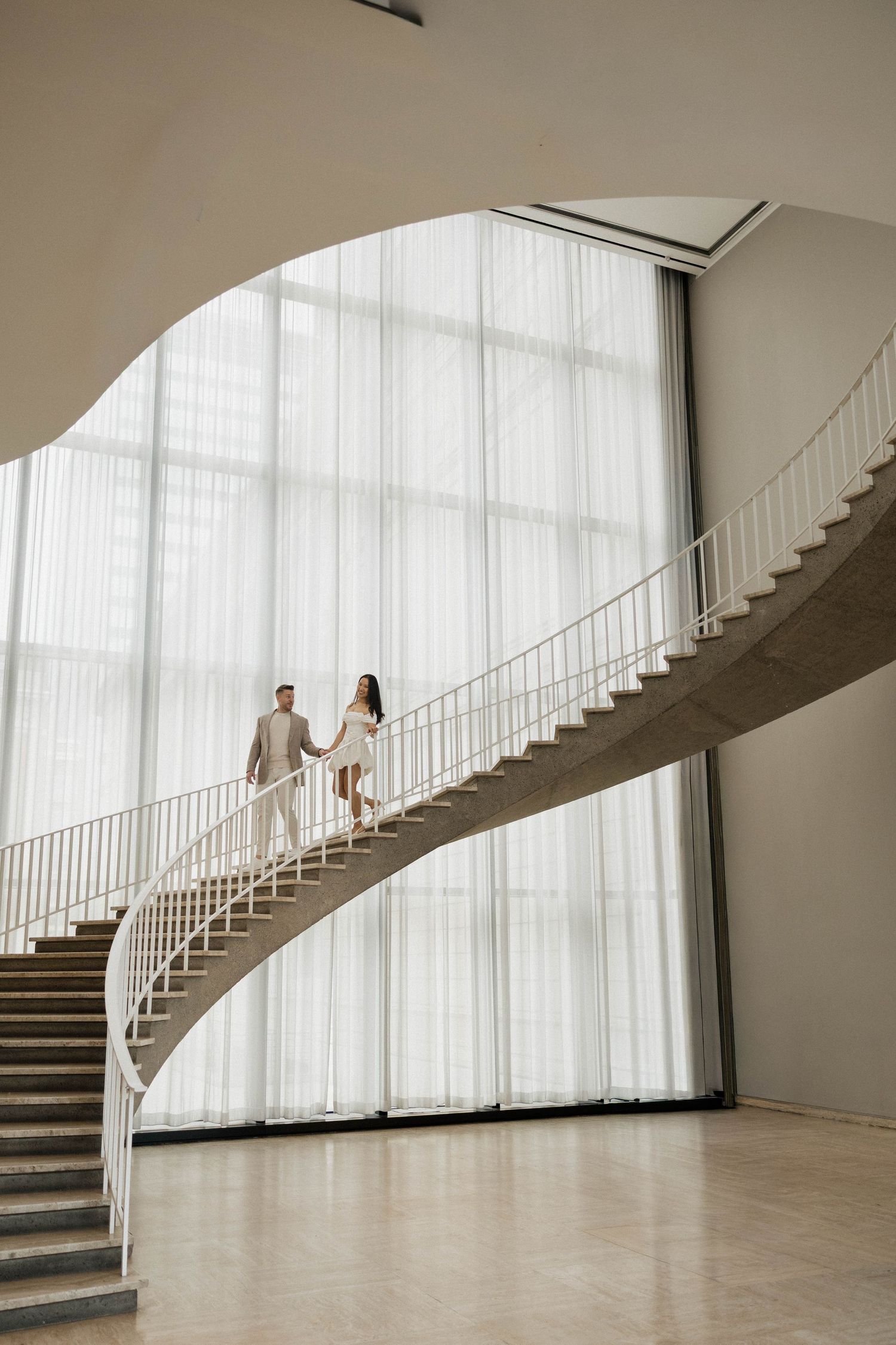Couple holding hands walking down elegant curved spiral staircase in bright modern building with floor-to-ceiling windows.