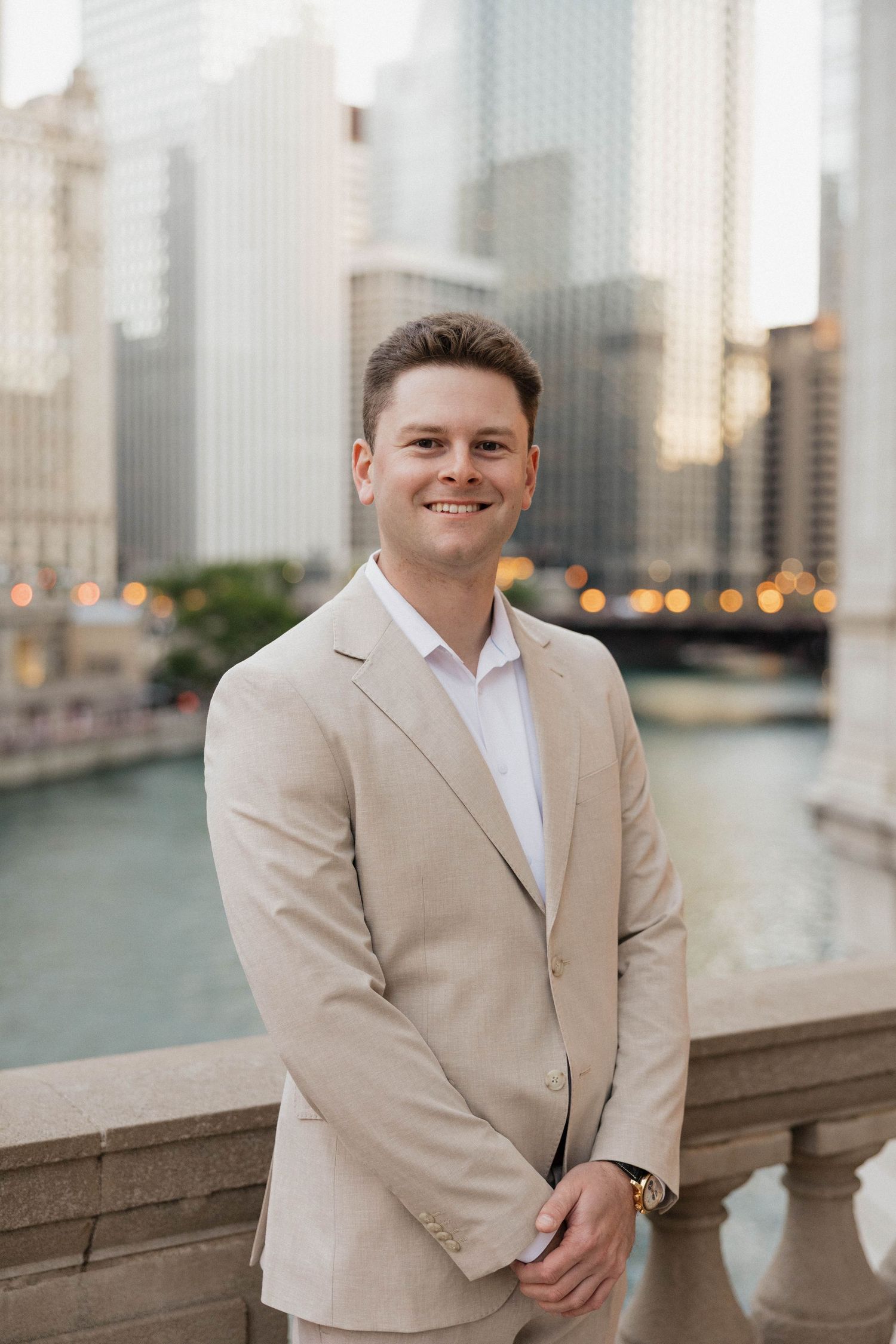 Young professional man in beige blazer smiling outdoors by Chicago River with city skyline in background.