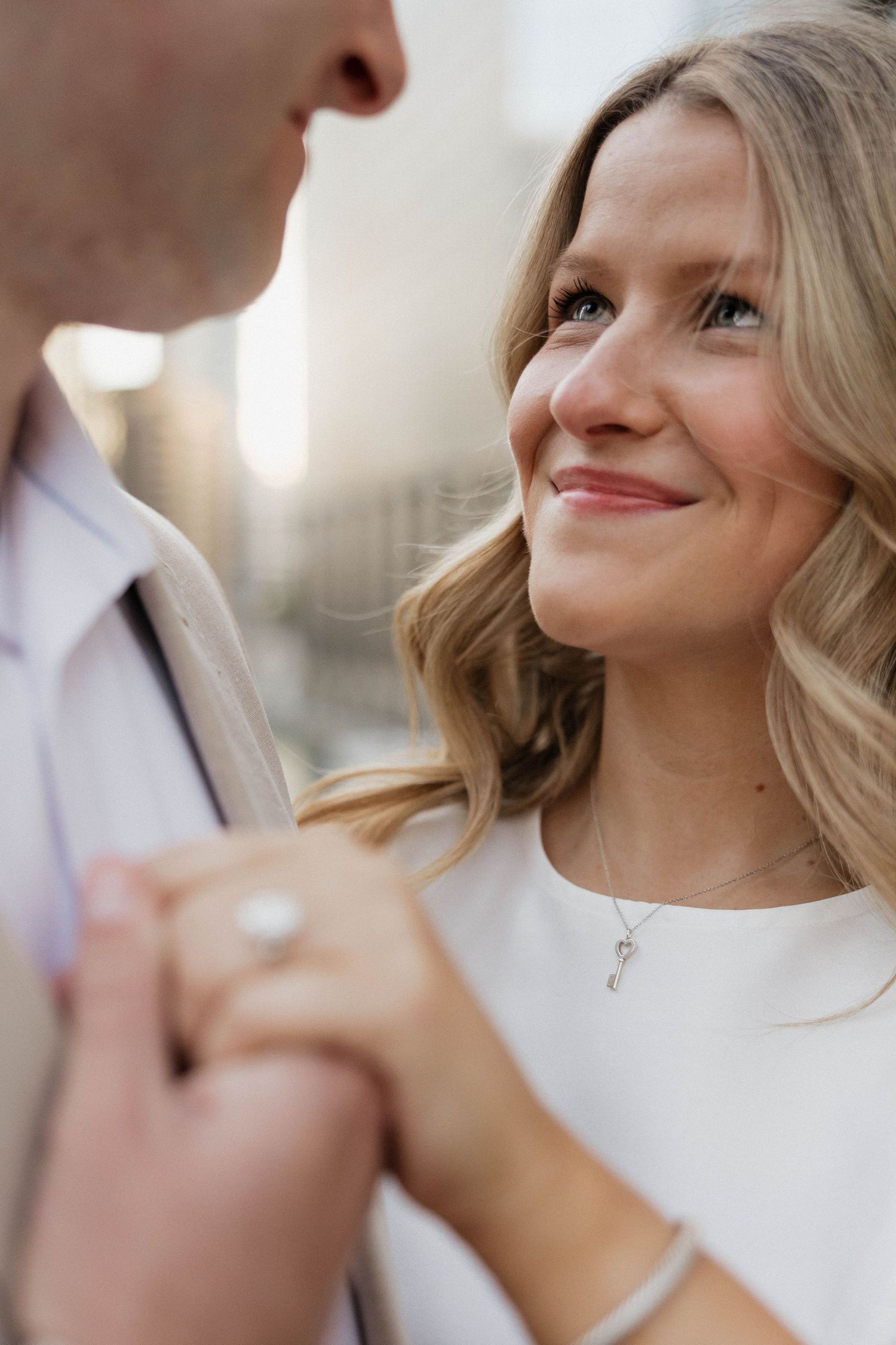 Smiling blonde woman gazes up at her partner while holding hands, wearing a white top and diamond ring.