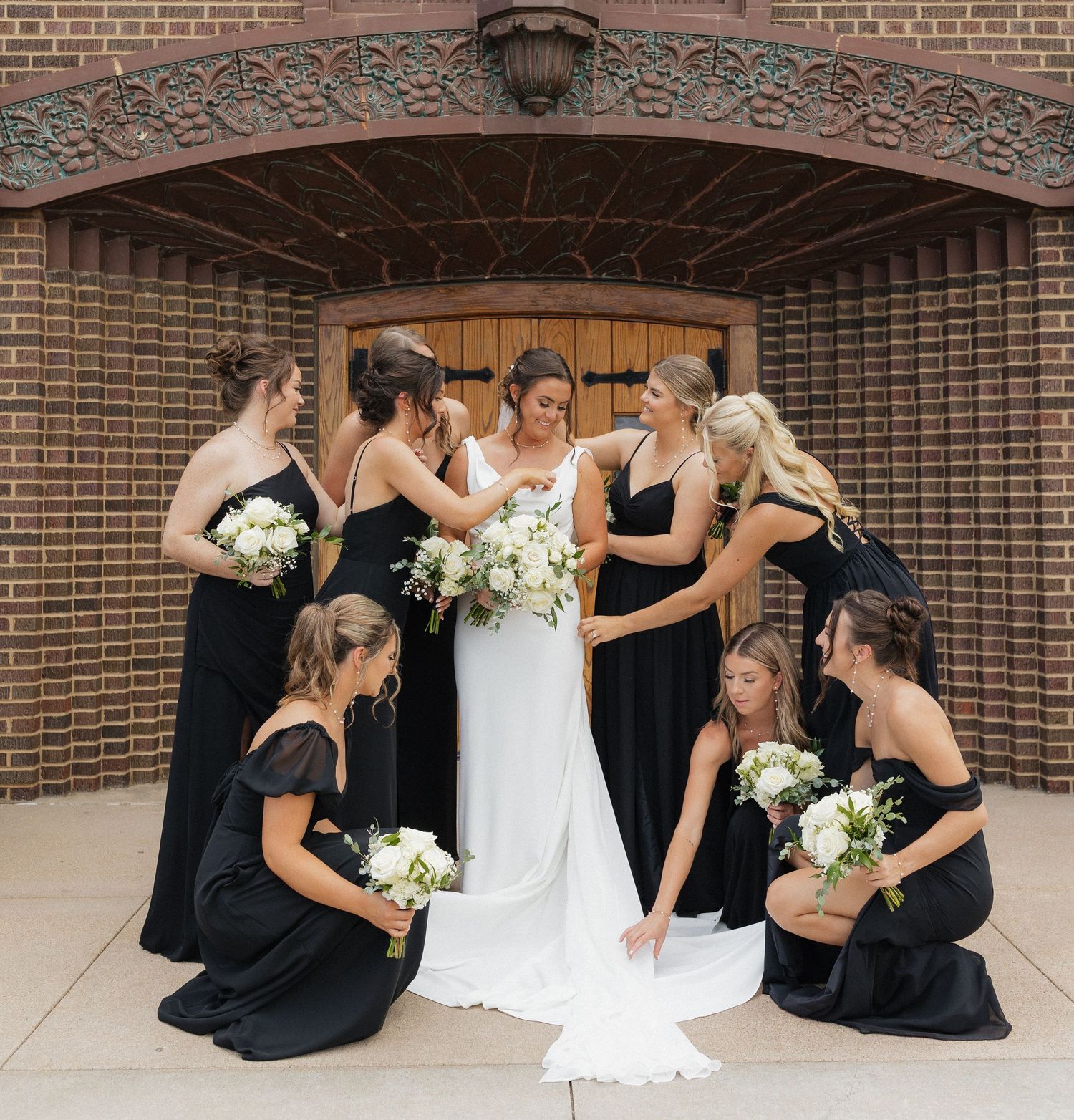 Bridesmaids in black dresses gather around a bride in white for candid photos on a brick patio.