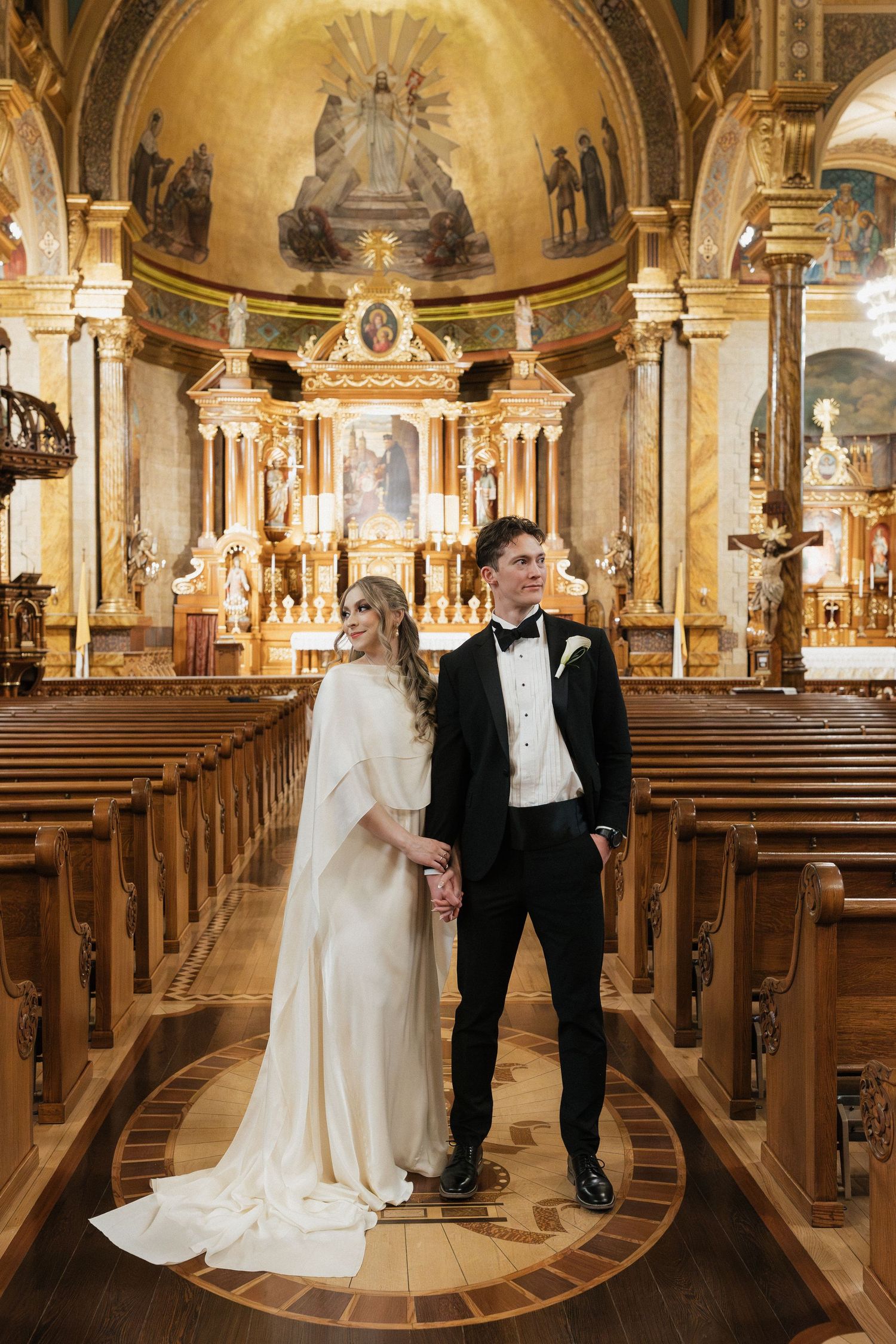 A couple stands in the aisle of a grand cathedral with ornate golden details and vaulted ceilings.