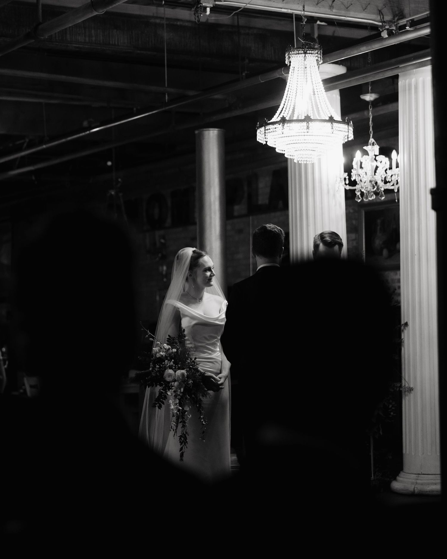 A black and white wedding scene with elegant crystal chandeliers and white columns in a dimly lit venue.