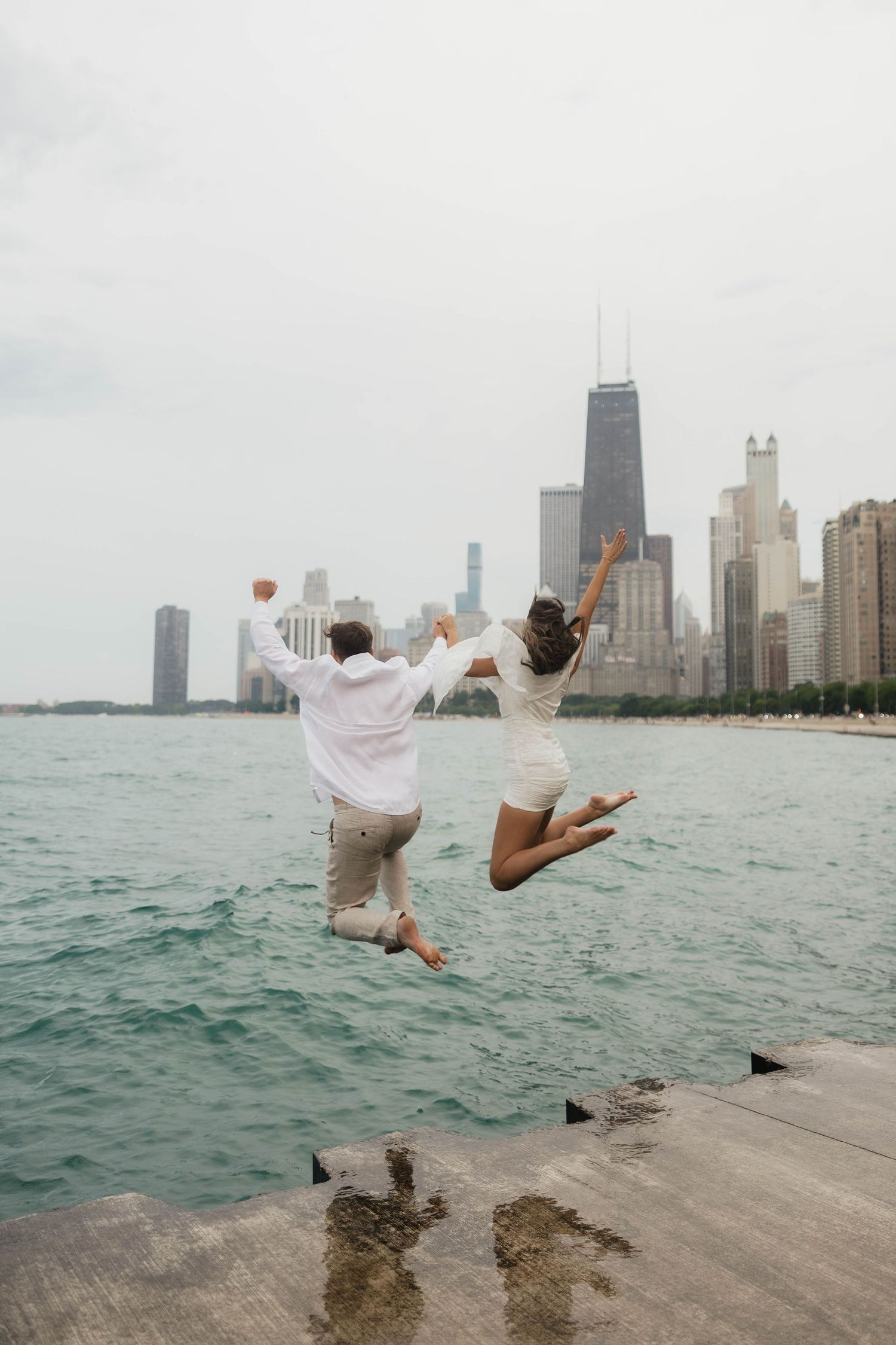 Couple jumping joyfully off a pier with Chicago skyline and Lake Michigan in the background on a cloudy day.