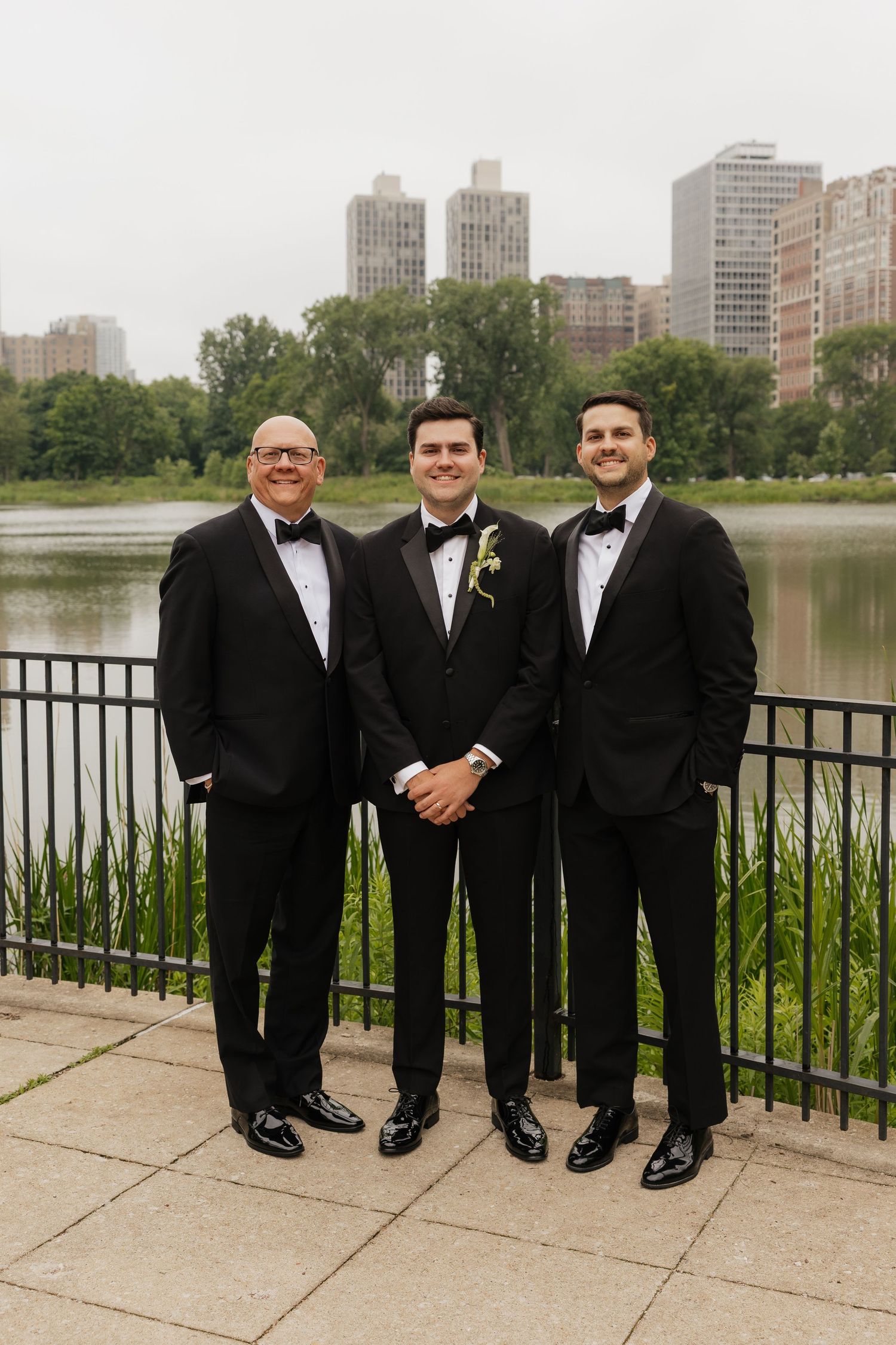 Three men in black tuxedos pose by a lakeside railing with city skyline and greenery in the background.