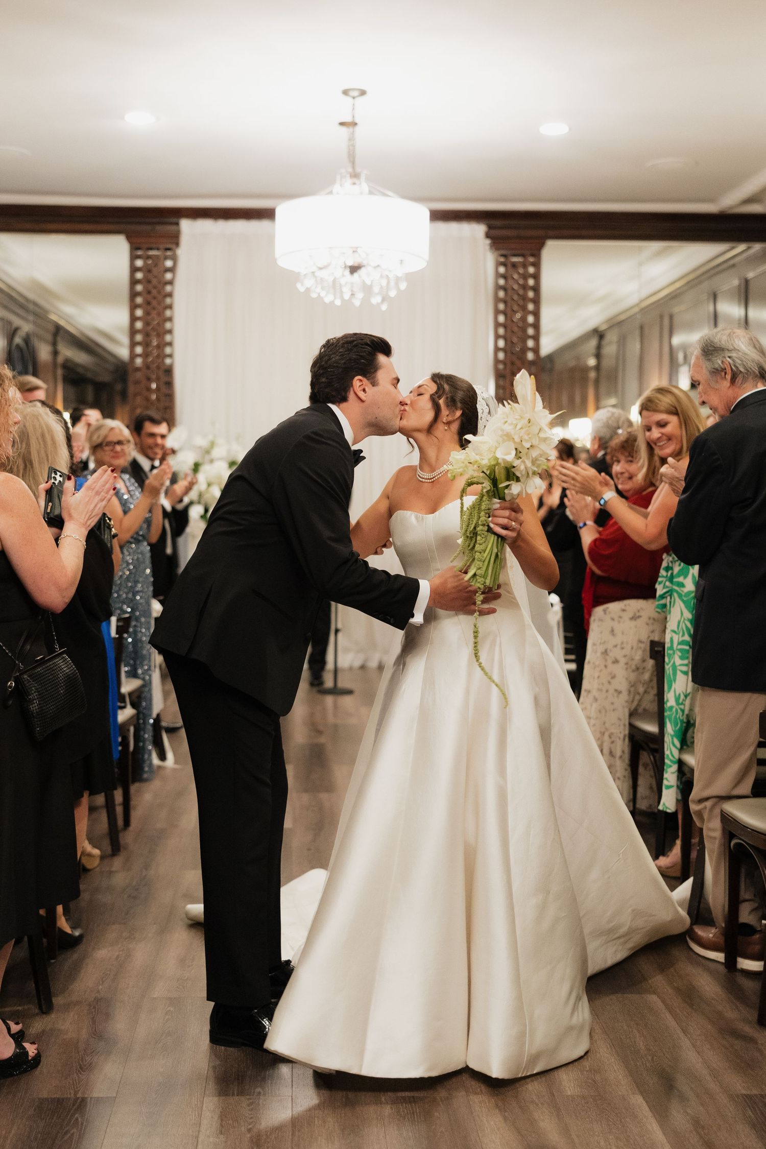 Newlyweds share a first kiss while walking down the aisle, bride holding white bouquet, guests applauding in elegant venue.