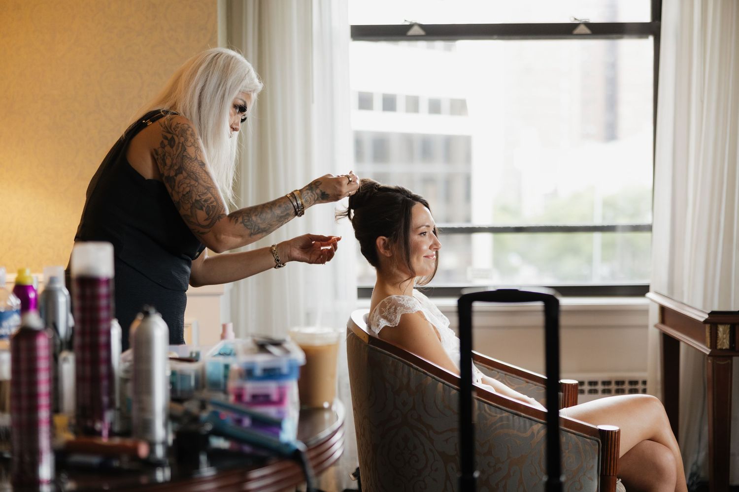 Hair stylist applying finishing touches to a smiling woman's updo in a hotel room with beauty products nearby.