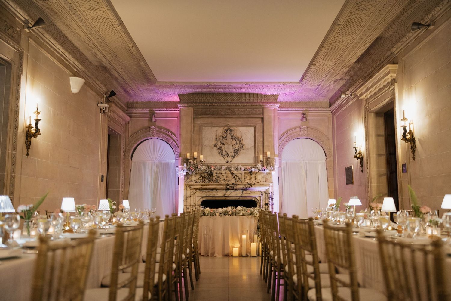 Elegant wedding reception hall with gold chairs, candlelit tables, ornate fireplace, and soft pink uplighting in a grand ballroom.
