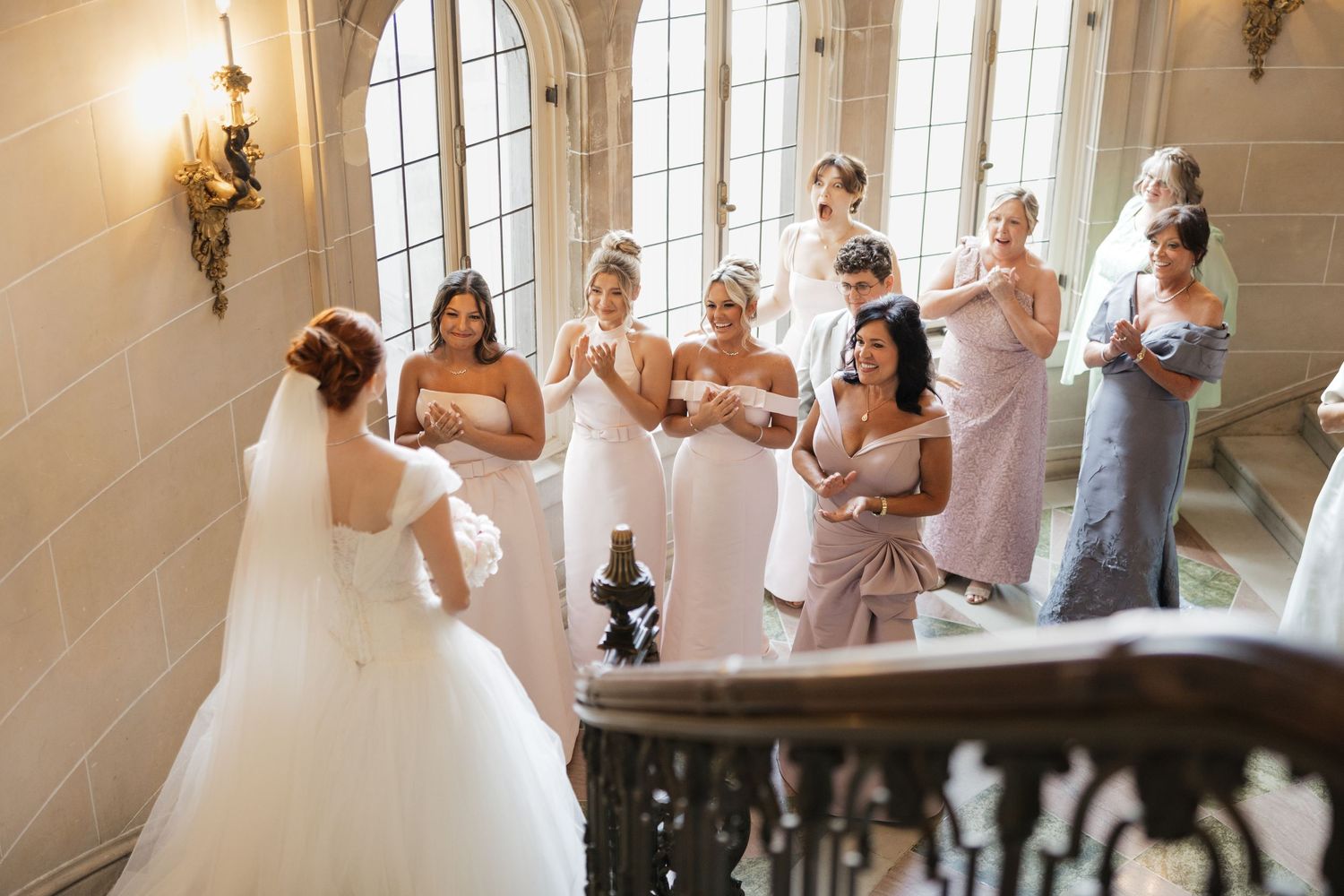 Bride in white gown descends staircase as bridesmaids in pink dresses react with joy at elegant wedding venue.