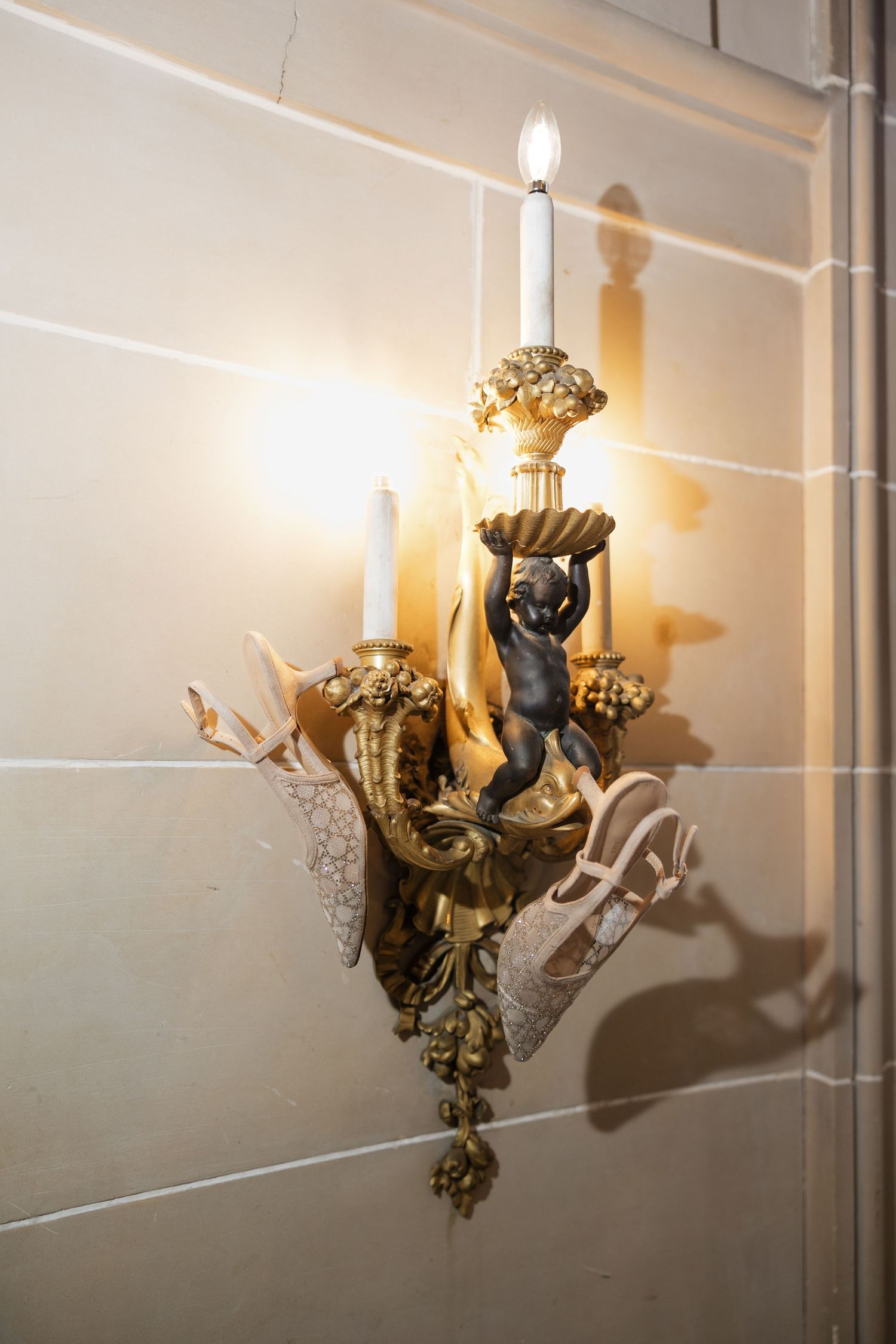 Ornate gold wall sconce with cherub figurine holding candelabra with lit candles mounted on tiled wall.