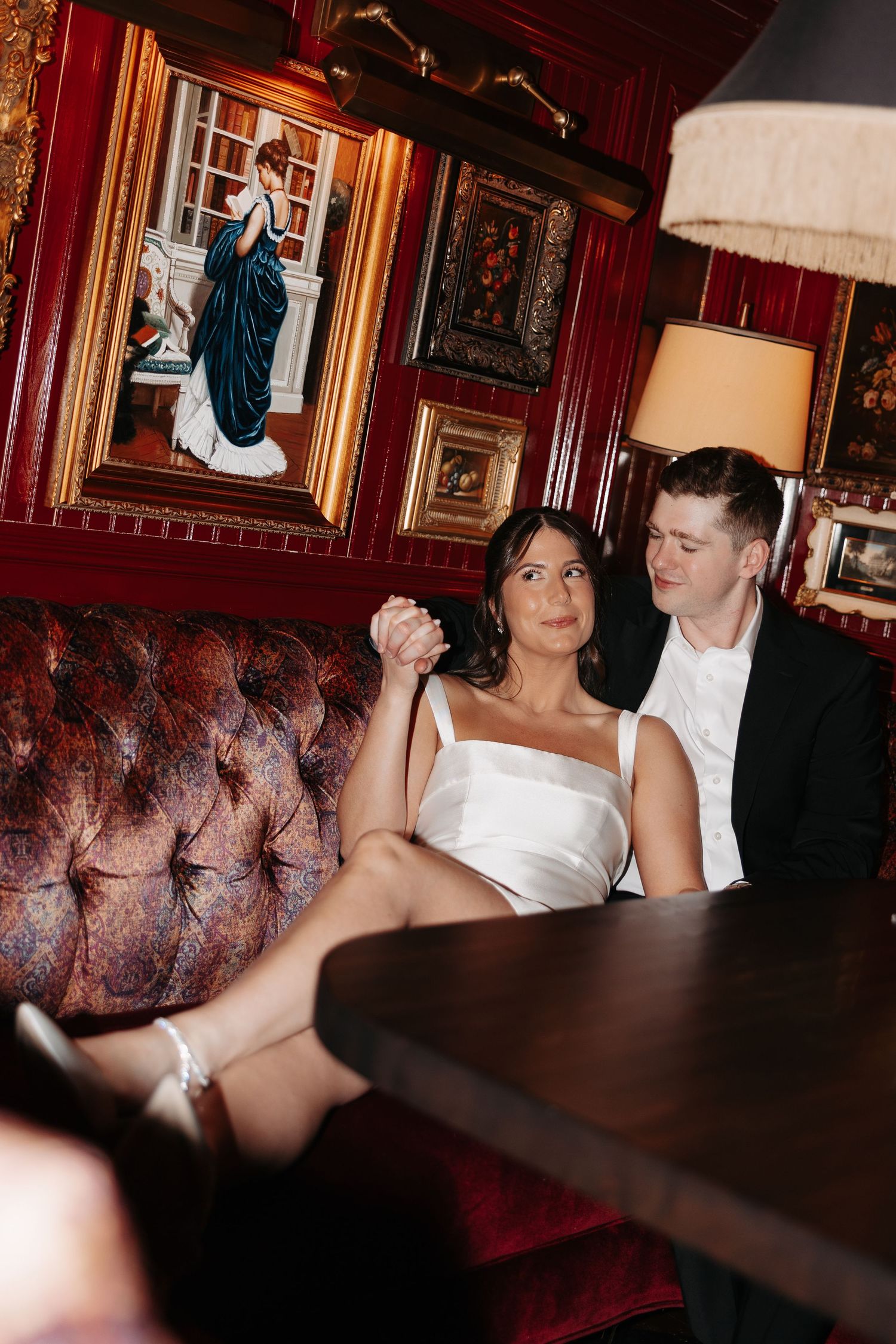 A couple shares an intimate dance moment on a brown leather couch in an elegant room with red walls and framed artwork.
