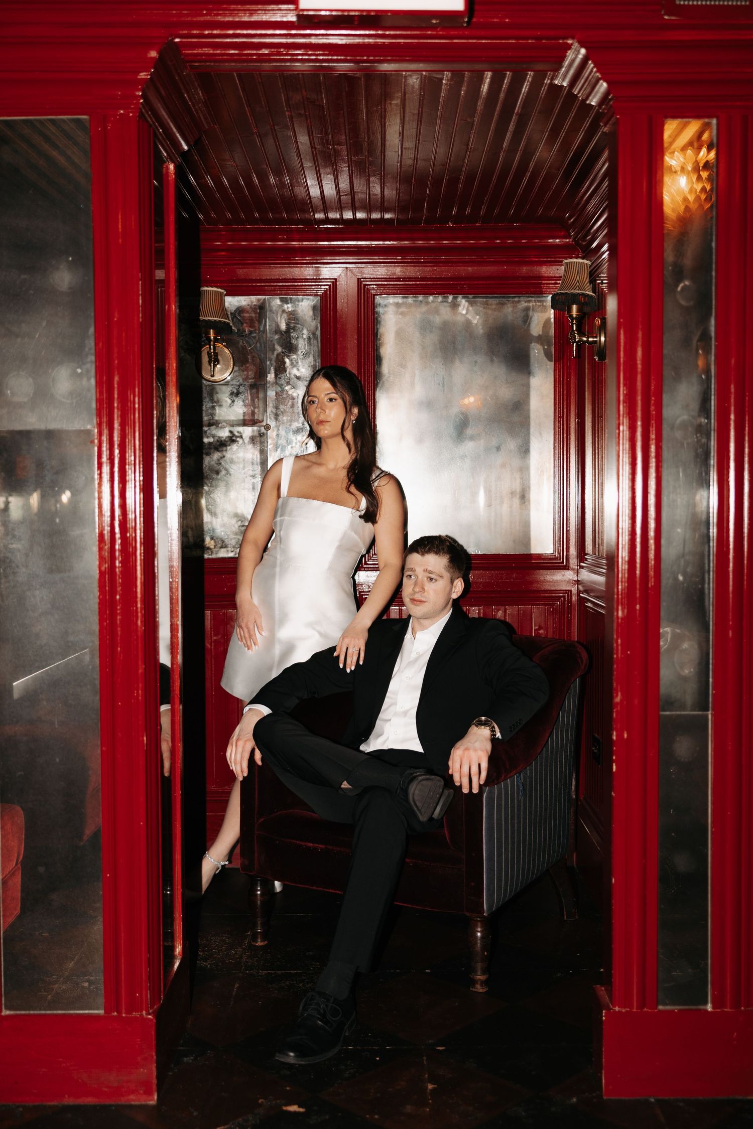 Dramatic fashion photoshoot in a red-walled corridor with moody lighting and formal attire.