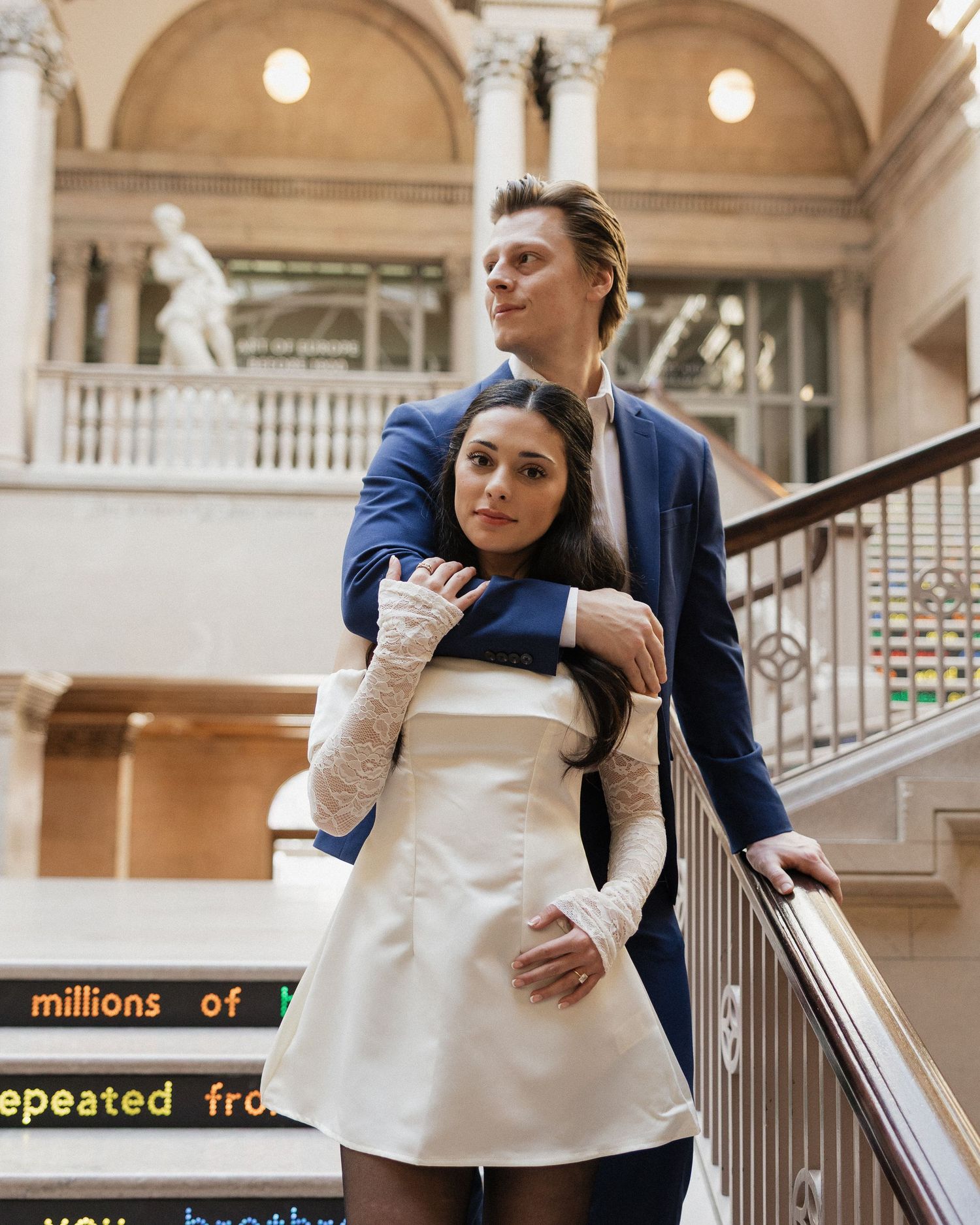 A couple embraces on an elegant marble staircase in a grand building with arched windows and classical architecture.
