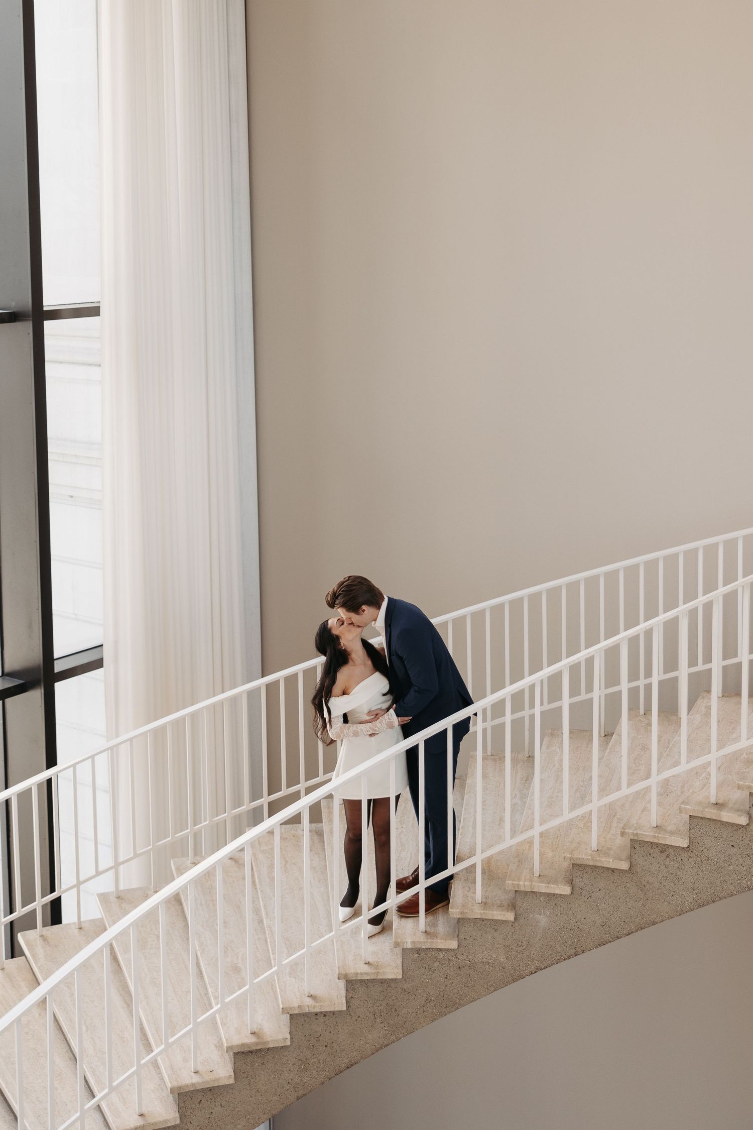 Couple sharing intimate moment on white marble staircase in formal attire.