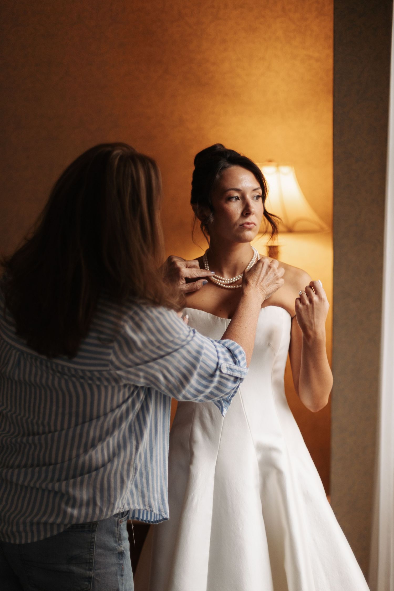Bride in a strapless white gown looks away as a woman in a striped shirt fastens her pearl necklace beside a softly lit lamp.