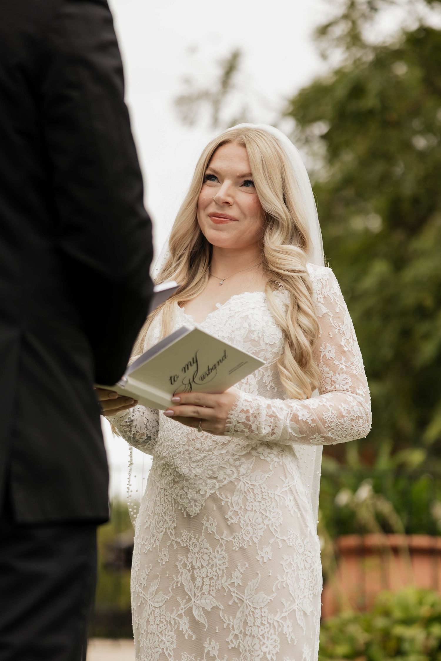 Smiling bride in a lace wedding gown and veil reads vows from a card labeled 