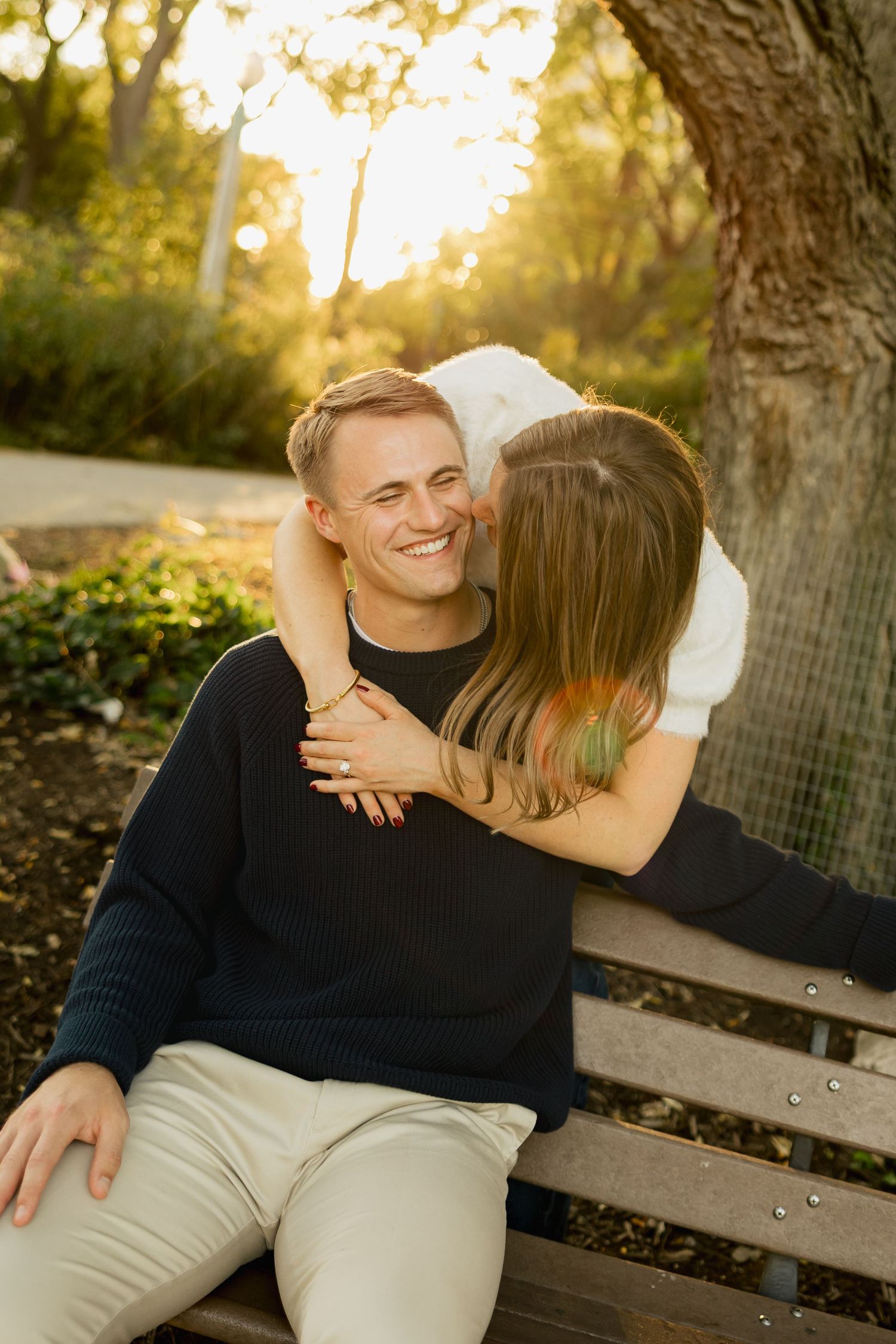 A smiling man in a navy sweater sits on a park bench as a woman with long hair hugs him from behind in warm golden sunlight.
