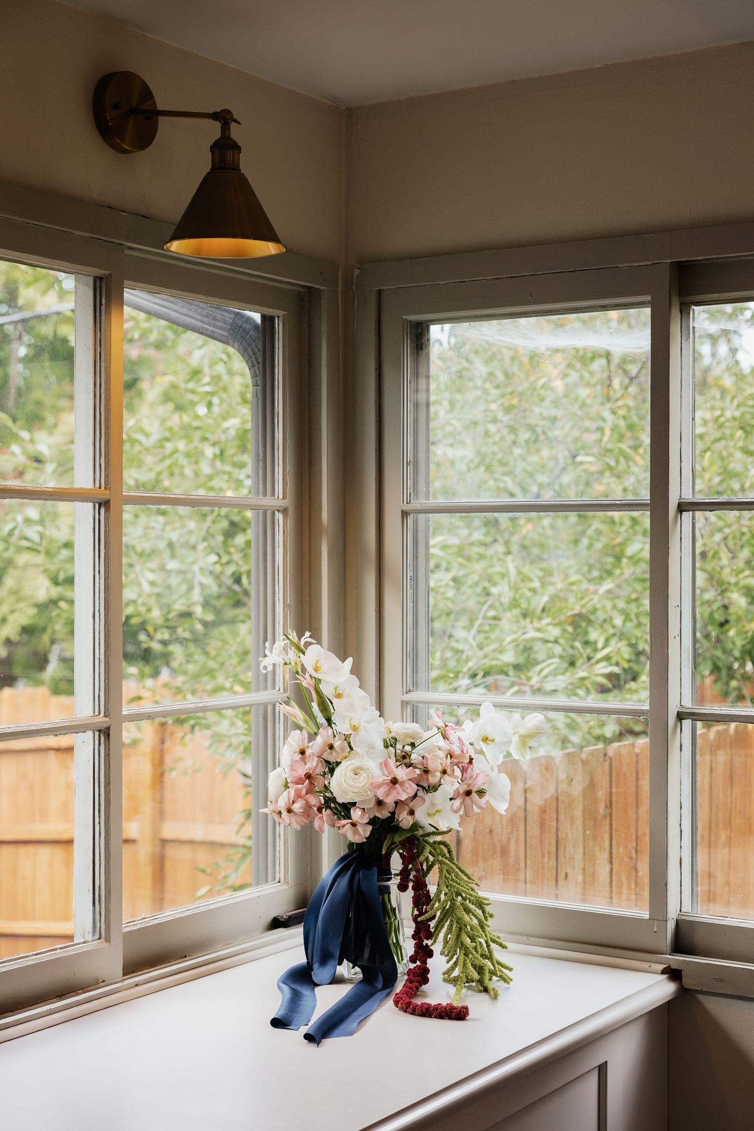 Bridal bouquet of pink and white flowers with a navy ribbon rests on a sunlit windowsill beside a vintage wall sconce.